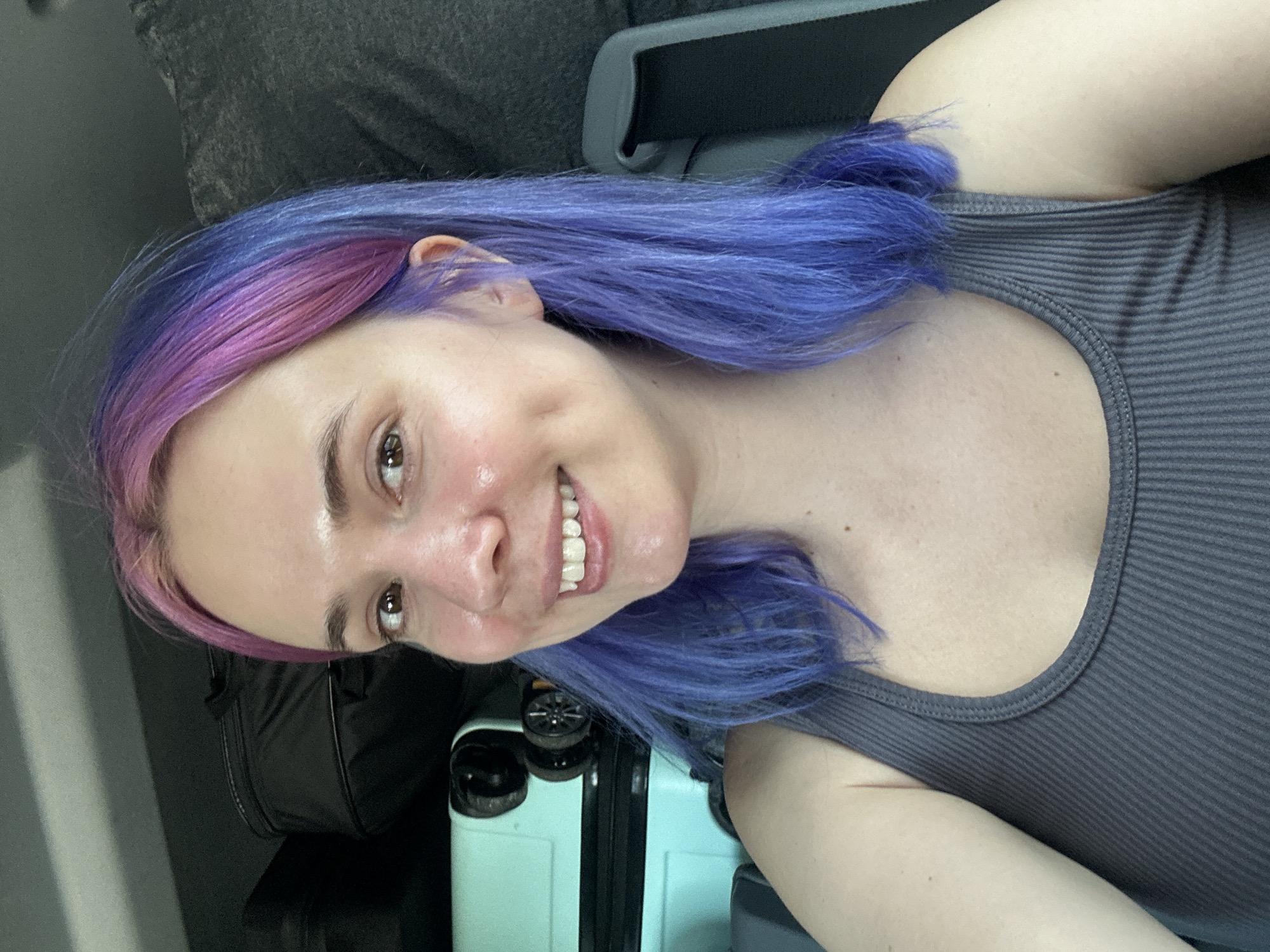 A young adult woman is pictured in a selfie, likely inside a vehicle. The subject has dyed hair in shades of pink and blue. She is wearing a gray tank top. A turquoise suitcase and a black bag are visible near her. The location is likely Obrežje, Slovenia. The image lacks specific landmarks or details to confirm this location definitively.