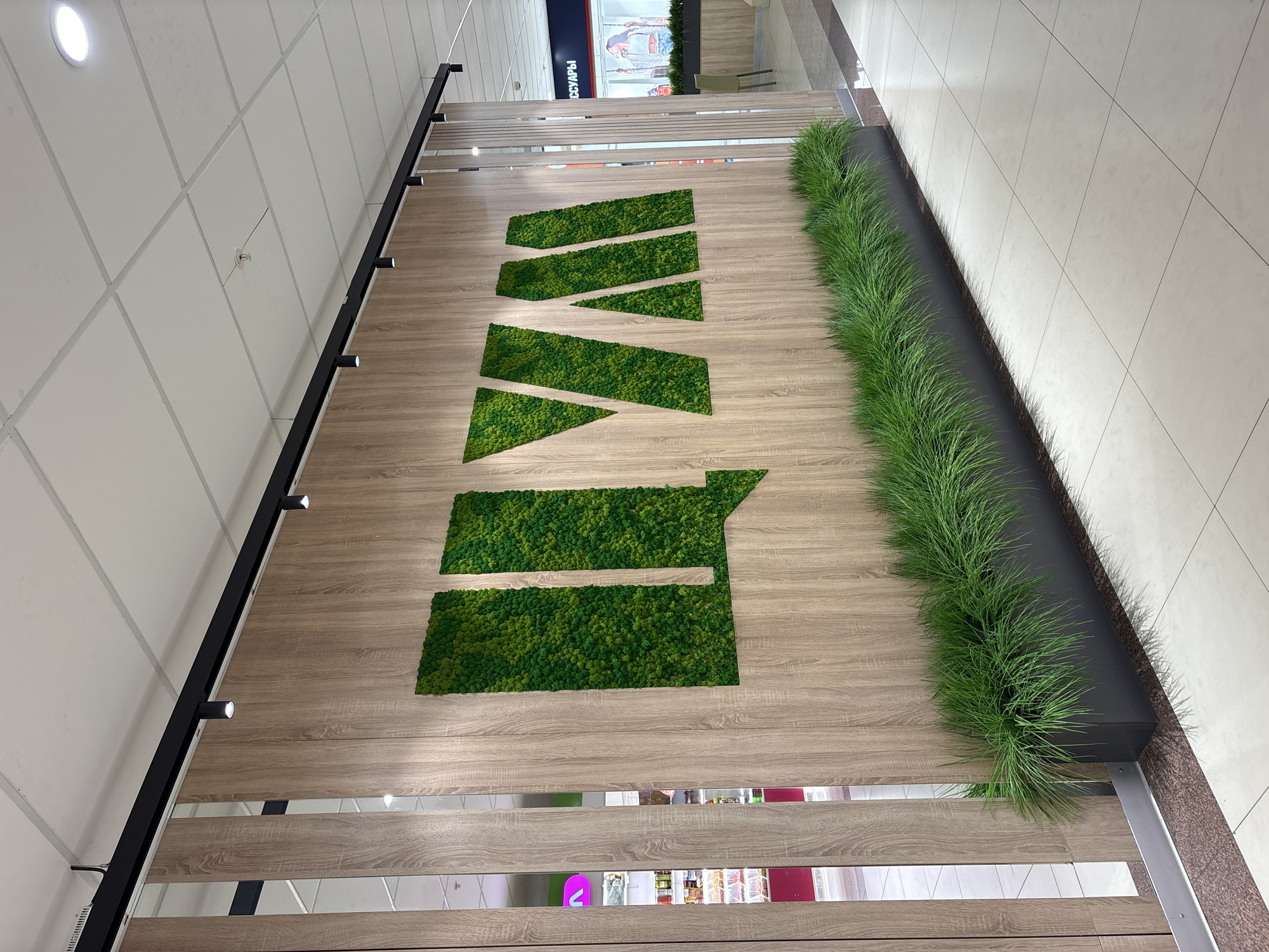 The image captures an interior wall display in an unknown location within the city of Unknown, Unknown. The display features a wooden panel with a company logo made of green moss in an abstract, linear style. A thick strip of long, thin faux grass runs along the upper right edge of the wooden panel, contrasting with the wood's texture and adding a natural element. The wall itself appears to be made of long, horizontal wooden planks with interspersed metallic, reflective accents. A black, track lighting fixture is installed along the top of the wall. The floor is composed of light-colored tiles. The scene is well-lit.