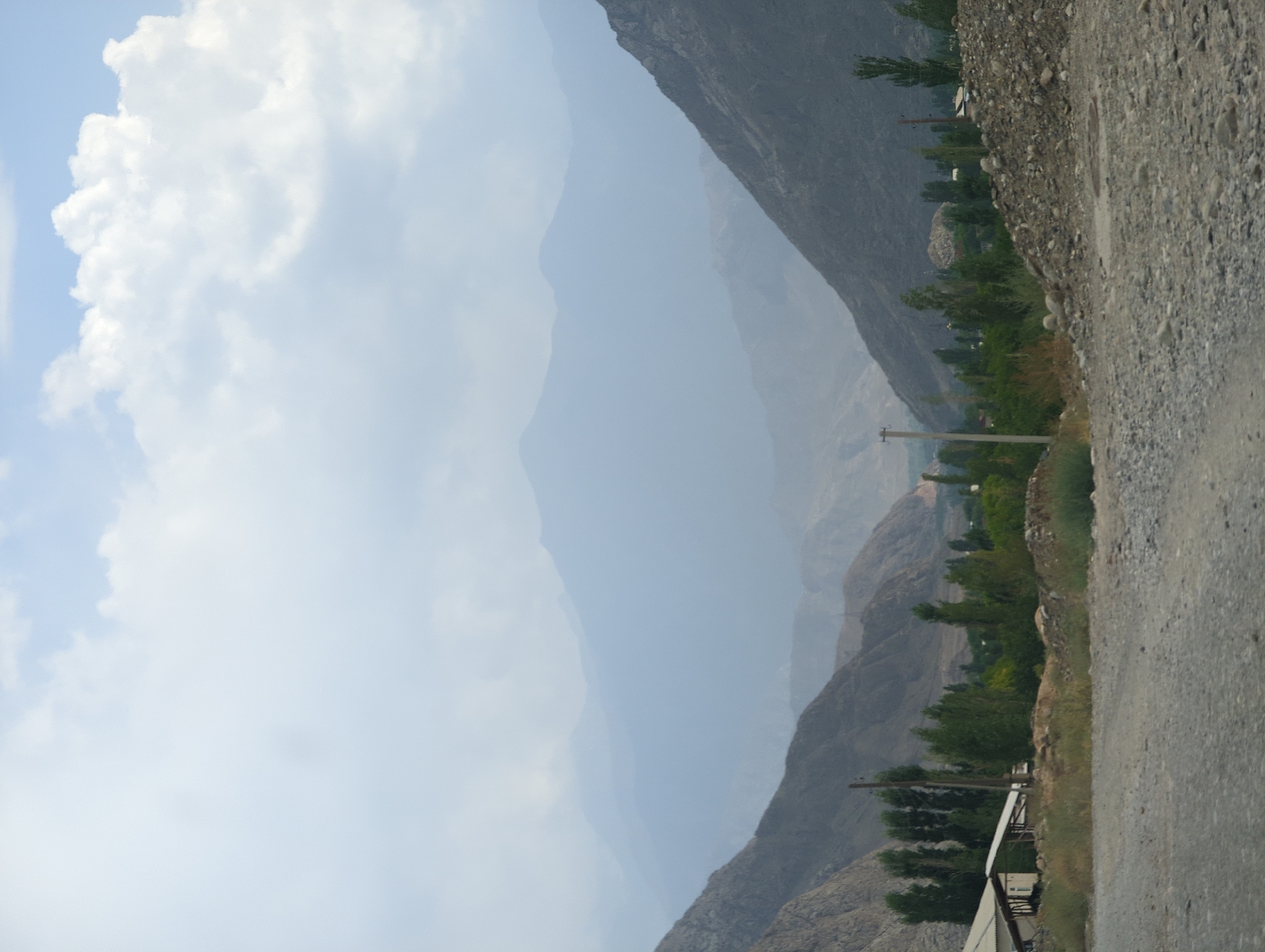 The image depicts a vast, mountainous landscape with a dirt road in the foreground. In the mid-ground, there is a small settlement with a few buildings and trees. The background is dominated by multiple layers of hazy mountains, suggesting a considerable distance and atmospheric conditions. The sky is partly cloudy, with patches of blue visible through the clouds. The overall scene conveys a sense of natural grandeur and remoteness.