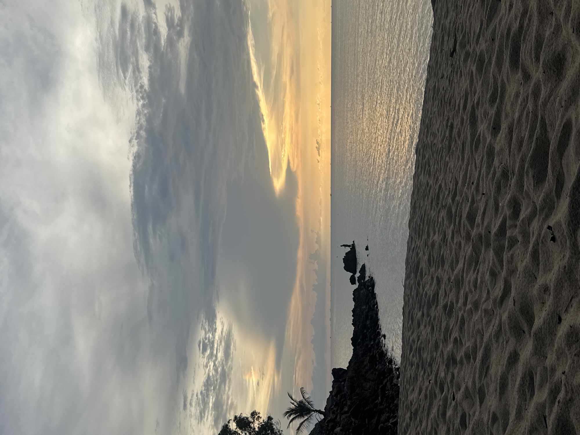 The image depicts a beach scene at Baan Wang Ta Kien, Thailand. The photo is oriented sideways. A sandy beach occupies the right side. Adjacent to the beach is the ocean, with calm waters reflecting the light. In the distance, there are rocky formations extending into the sea with two people standing on the rocks. Above the ocean, the sky is filled with clouds, showing a mix of grays, whites, and golden hues indicating either sunrise or sunset. Some trees are visible near the rocks along the shoreline.