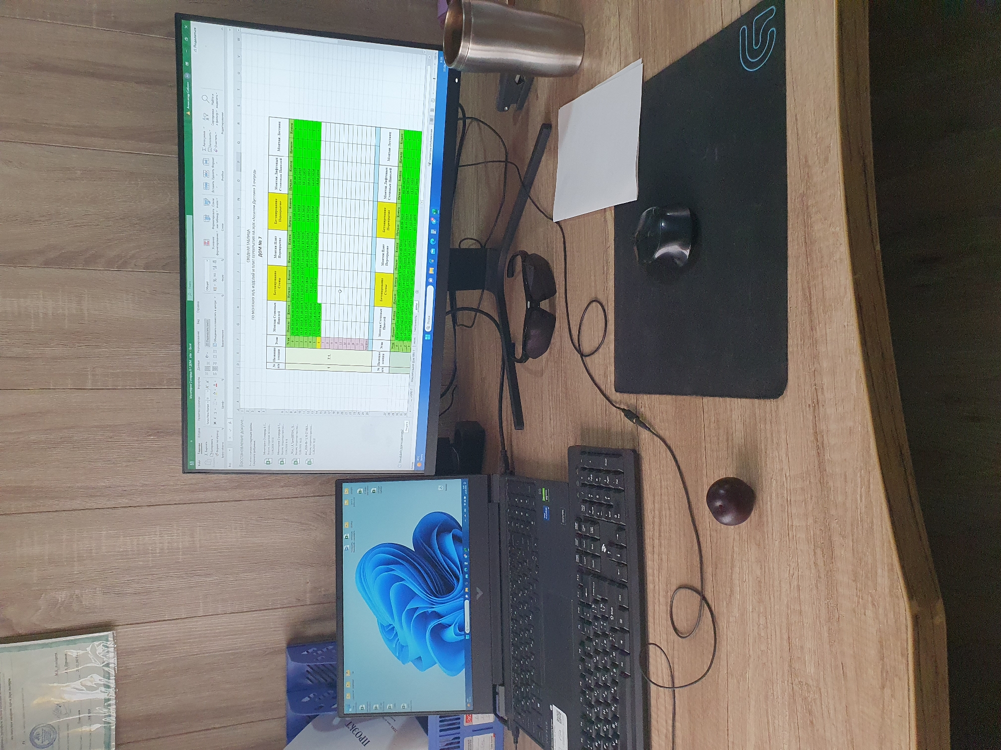 The image depicts a desk setup with a laptop, an external monitor displaying a spreadsheet, a keyboard, a mouse, sunglasses, a metal tumbler, and a plum. The laptop screen shows the Windows 11 operating system. The external monitor displays an Excel spreadsheet with columns for "№ п/п", "Этаж", "Монтаж Стенных Панелей", "Бетонирование перекрытий", "Монтаж плит перекрытий", "Бетонирование перекрытий", "Монтаж лифтовых Шахт", and "Монтаж лестниц", with rows containing dates and numerical data. The spreadsheet's title appears to be "СВОДНАЯ ТАБЛИЦА ПО МОНТАЖУ Ж/Б ИЗДЕЛИЙ И ПРИ ТЕРРИТОРИЯХ НА ЖК «Асалом Дустлик» 3 очередь ДОМ №7". The background consists of a wood-paneled wall. Based on the language and content of the spreadsheet, the scene is likely an office or workspace in Uzbekistan. While a specific location in Tashkent cannot be determined from the image alone, the presence of Cyrillic script and the context of construction/engineering data suggest a professional setting within the country. There are no people visible in the image, and no actions are being performed by any subjects. The environment is a static desk setup.