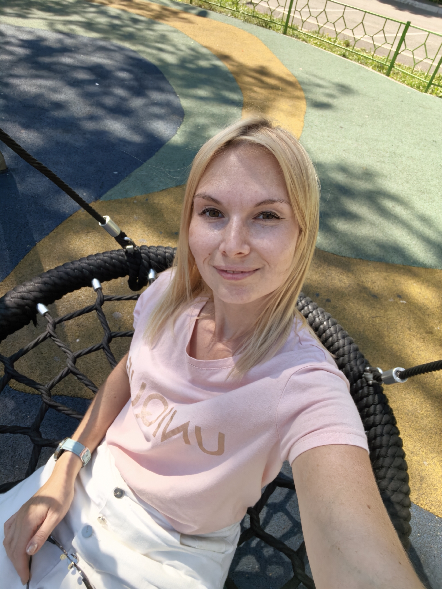 A blonde woman with fair skin and freckles is seated on a round, net-like swing made of black rope. She is wearing a light pink t-shirt with gold lettering and white pants. A light blue watch is visible on her left wrist. The swing is suspended by thick black ropes. The ground beneath her is a colorful, patterned rubberized surface in shades of blue, yellow, and green. In the background, a green metal fence with a geometric pattern is visible, with grass and a road beyond. The scene appears to be an outdoor playground. The image does not contain any information linking it to the city of Sterlitamak, Russia.