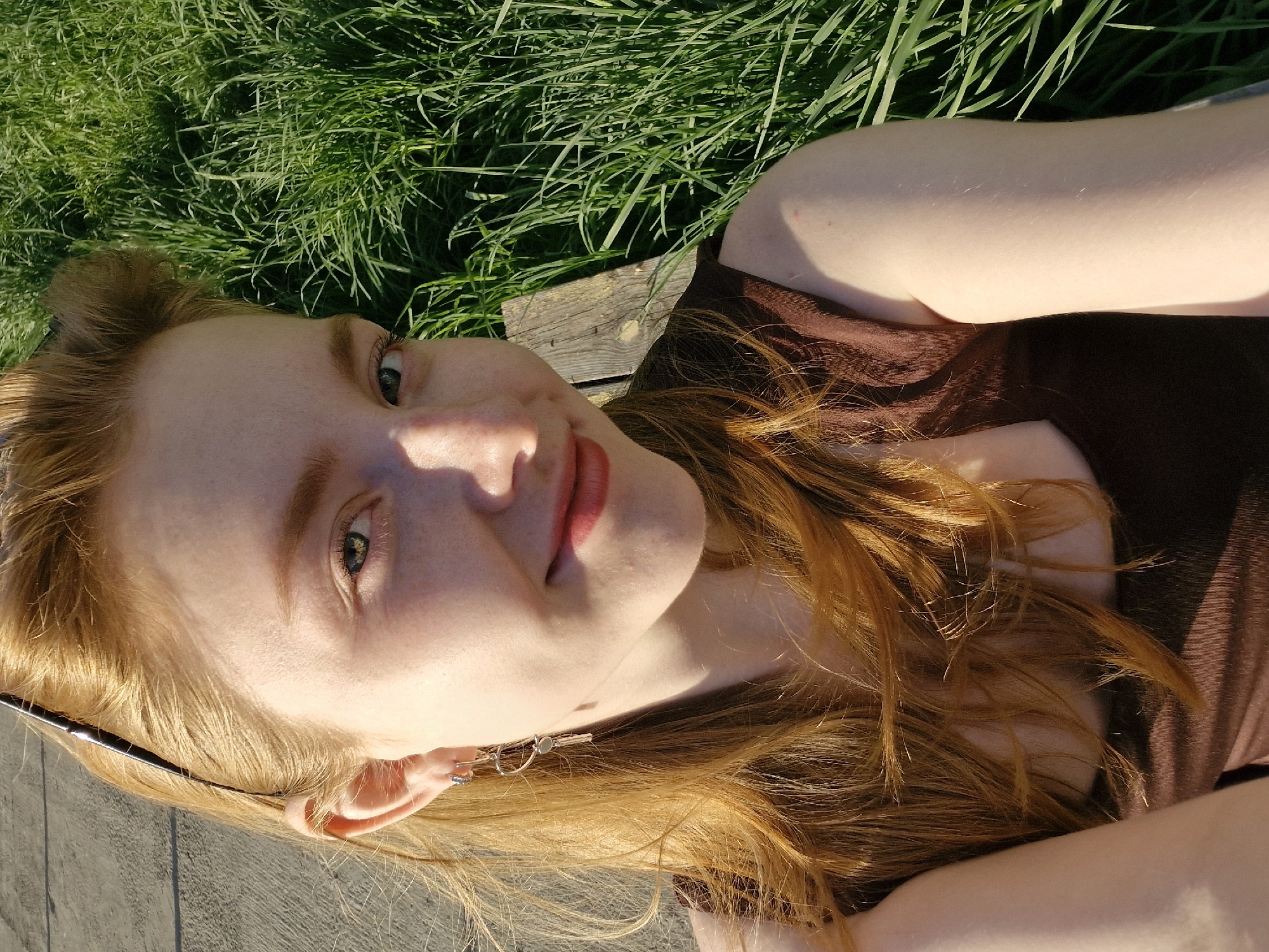 A young, fair-skinned woman with red-golden hair is lying on what appears to be a wooden bench or platform. She is wearing a brown top and has earrings. Lush green grass is visible behind the bench. The overall scene suggests a sunny day in an outdoor setting, likely a park or recreational area. The location is specified as Yekaterinburg, Russia.