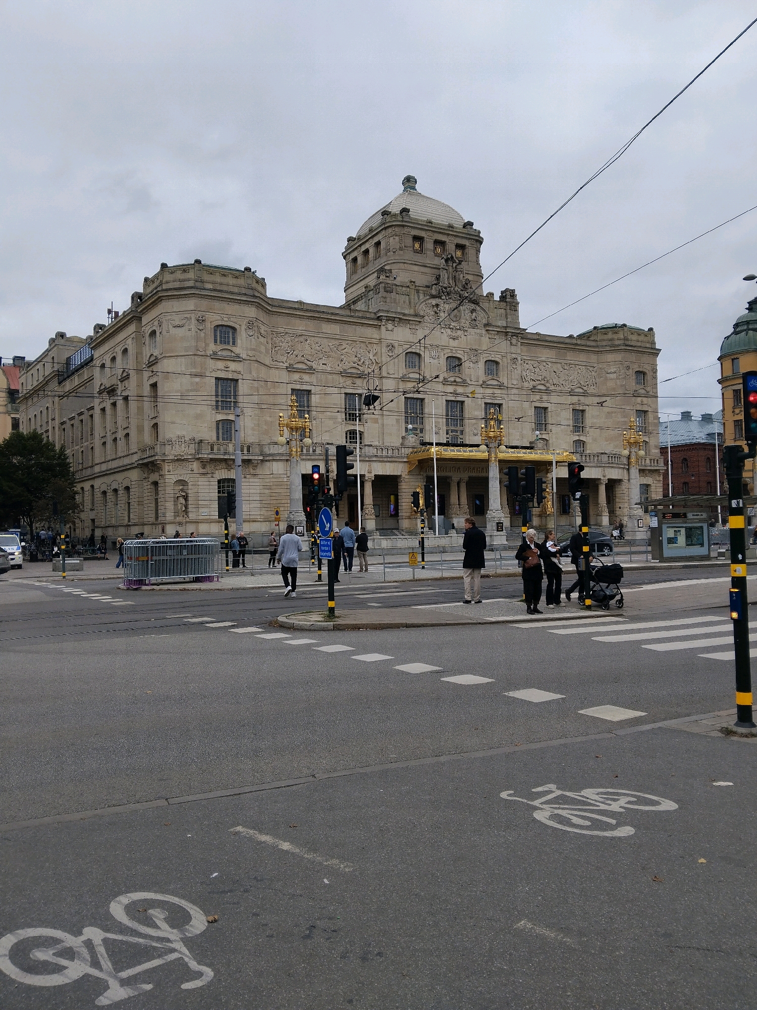 The image depicts the Royal Swedish Opera house in Stockholm, Sweden, on an overcast day. The grand building, constructed of light-colored stone, features intricate architectural details, a prominent dome, and gilded decorative elements above the entrance. In the foreground, a city street is visible with a pedestrian crossing and a marked bicycle lane. Several people are crossing the street and waiting at the intersection, including a person pushing a stroller. Traffic lights are red for vehicles, and a blue traffic sign with a white arrow indicates a direction. A temporary metal barrier is positioned on the sidewalk. The street is paved, and there are some scattered leaves on the ground. The overall atmosphere is urban and functional.