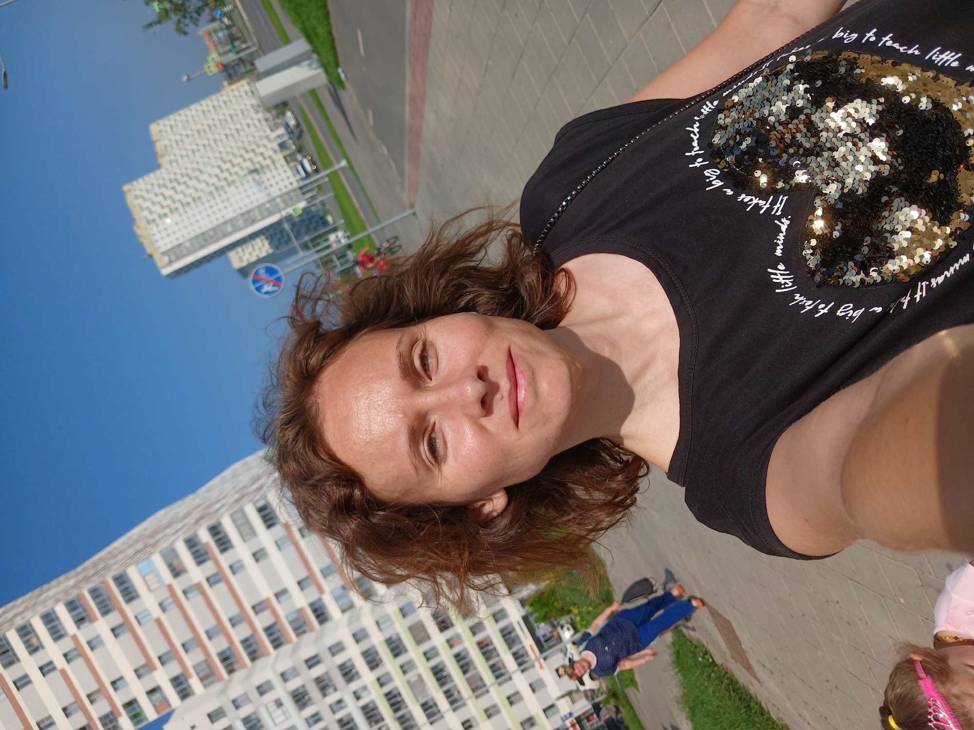 A woman is taking a selfie outdoors in front of tall apartment buildings in Yekaterinburg, Russia. She has brown, curly hair and is wearing a black t-shirt with a sequined heart design and text that reads "It takes a big to teach little minds." The background shows modern, high-rise buildings with balconies and windows, a paved walkway, green grass, and a street with some cars. In the background, another woman in a blue shirt and pants is walking away from the camera, and a child with a pink crown is partially visible in the foreground. The sky is clear and blue.