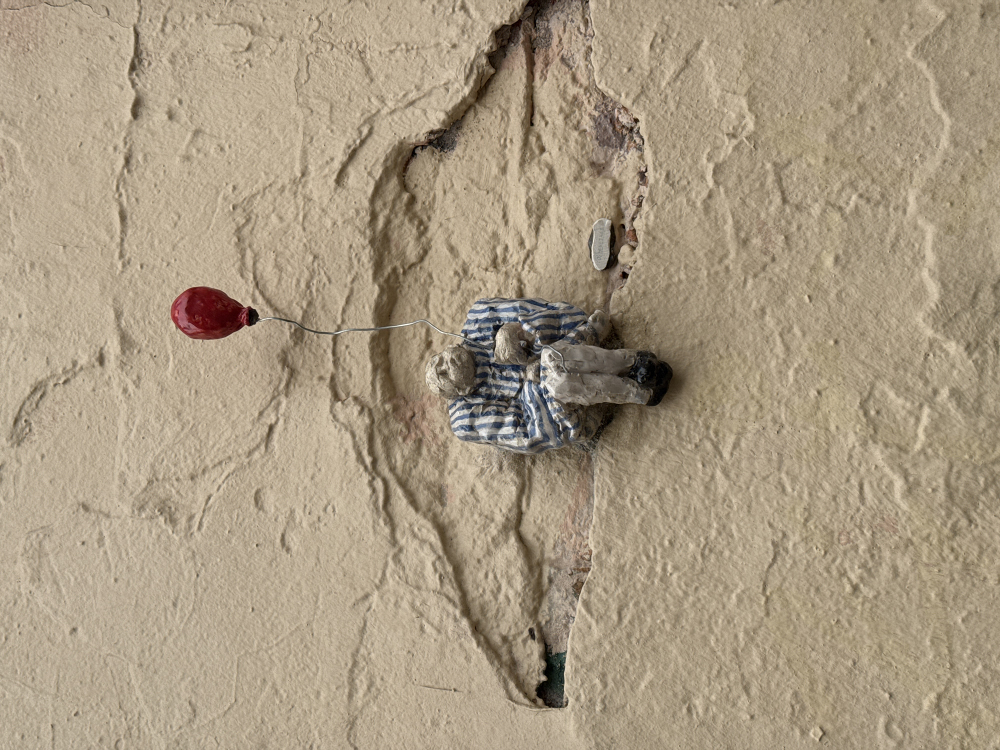 A miniature sculpture of a person wearing a striped shirt and pants sits on a textured, pale yellow wall in Saint Petersburg, Russia. The figure is seated and appears to be holding a small object. A red balloon is attached to the figure by a thin wire, floating above it. The wall has a rough, uneven surface with visible cracks and indentations. A small, oval-shaped plaque with text is affixed to the wall near the figure.