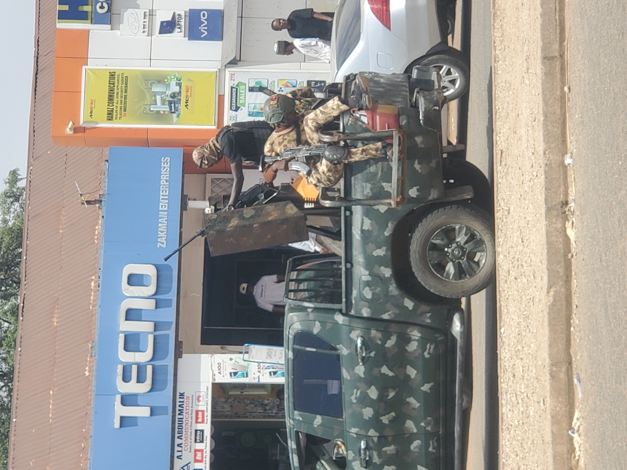 The image depicts a military vehicle, likely a pickup truck, camouflaged with green, white, and gray patterns. Two armed soldiers are in the back of the vehicle, one standing with a rifle and another seated with a rifle. The vehicle is parked on a street in front of a storefront displaying the name "TECNO" and the slogan "ZAKMAN ENTERPRISES." Another sign reads "ALA ABDULMALIK COMMUNICATION." Additional signage includes "HAMAZ COMMUNICATIONS, DEALER OF ALL HOME APPLIANCES, TELECOMS AND SECURITY GADGETS." Other stores displaying Vivo products are visible. The presence of armed soldiers and military equipment in front of a commercial area suggests a security or patrol operation, possibly in Jos, Nigeria.