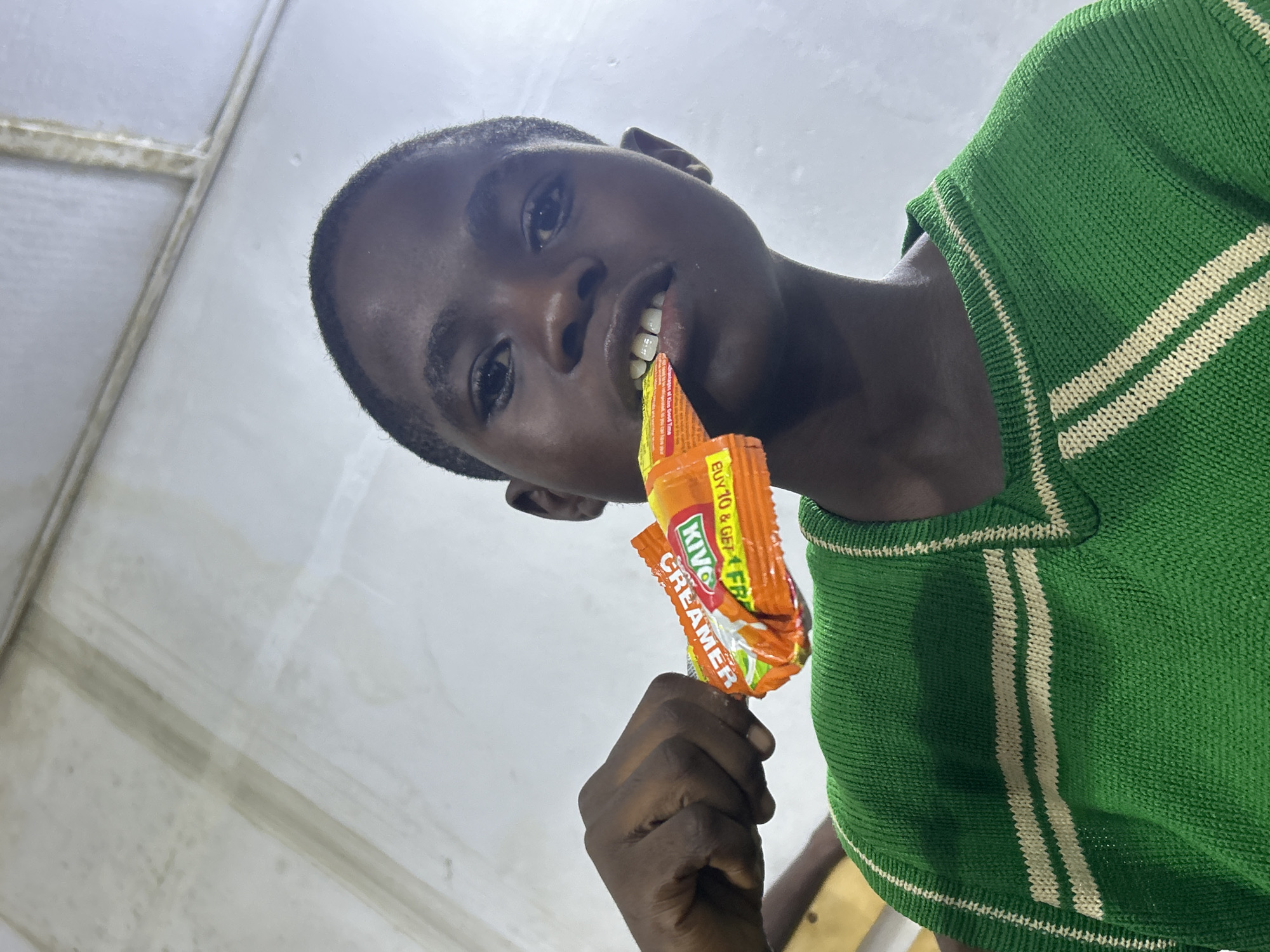 A young man is holding a small packet of Kivo creamer between his teeth and in his left hand. He is wearing a green knitted sweater with white stripes and standing indoors. The location is the city of Unknown, Unknown.