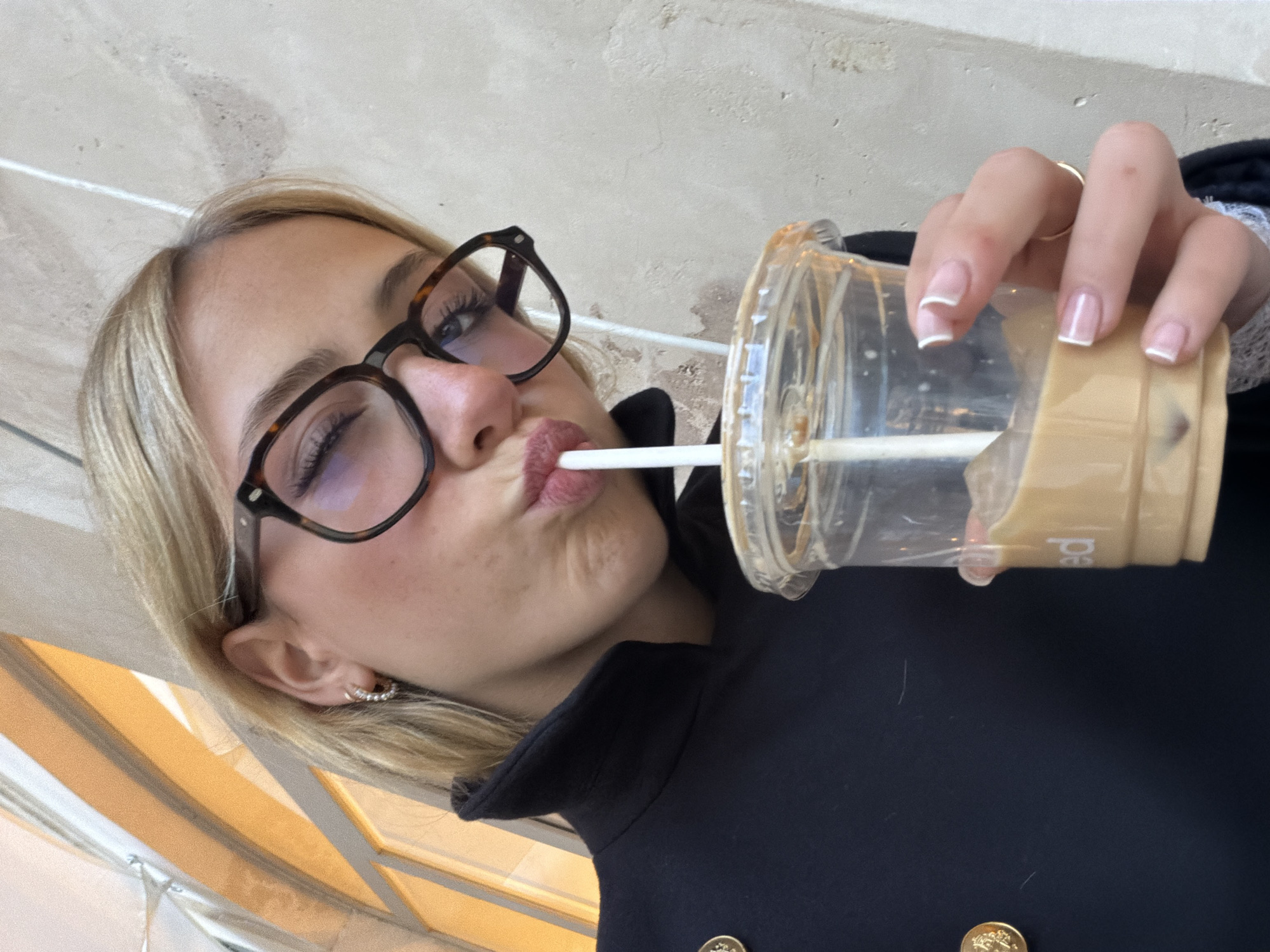 A blonde-haired individual, wearing tortoise-shell eyeglasses and a dark collared jacket with a gold button, is depicted in a close-up selfie. The individual winks with their left eye and puckers their lips around a white straw, actively drinking from a clear plastic cup containing an iced, light brown beverage. Their right hand, adorned with a gold ring and French-tipped fingernails, holds the cup. A small, textured hoop earring is visible in the individual's left ear. The background consists of a light-colored, textured wall and, to the left, an illuminated arched architectural structure with warm, yellowish light, indicative of an arcade or covered gallery in Paris, France.