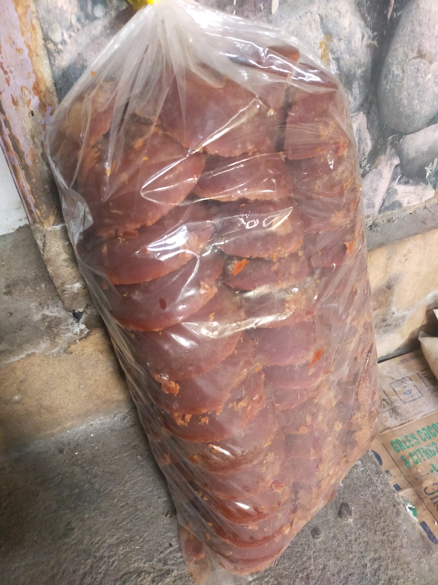 A clear plastic bag, filled with numerous circular, reddish-brown, flattened discs, rests on a concrete floor in Mojodanu, Indonesia. The discs appear to be palm sugar or a similar confection. The bag is positioned near a wall with textured, weathered paint and possibly some stone elements visible. To the right of the bag, a portion of a cardboard box with green text is visible. The lighting suggests an indoor environment, possibly a market or storage area, with diffuse light casting soft shadows. No people are present in the image.