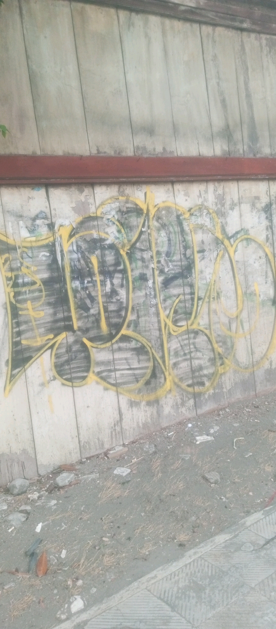 The image shows a close-up of graffiti on a weathered wall in Kediri City, Indonesia. The graffiti is in yellow lettering with black outlines, and the words appear to be "FJBO". The wall is made of vertical panels and is stained and worn. The ground in front of the wall is covered in dirt, debris, and some sparse dry grass. There is a red metal beam running horizontally across the top of the graffiti. The overall scene suggests an urban, possibly neglected, outdoor environment.