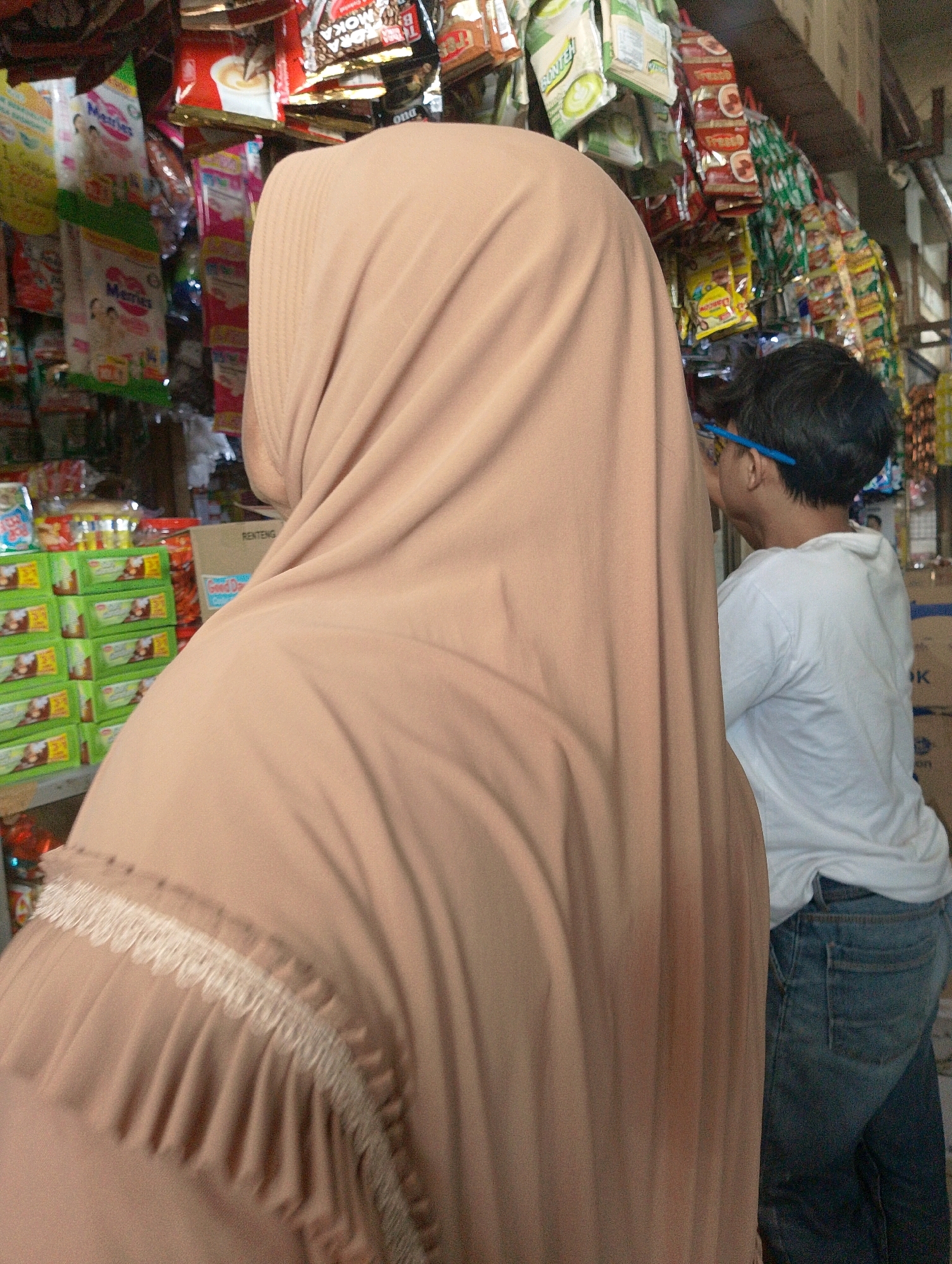 The image depicts a market stall in Lembang, Indonesia, filled with various packaged food items, primarily snacks and instant beverages, hanging from the ceiling and displayed on shelves. A person wearing a light brown hijab is in the foreground, facing away from the camera and towards the stall. To the right of the person in the hijab, a young male wearing a white t-shirt and blue jeans is partially visible, also facing the stall and appearing to be browsing or selecting items. The stall is crowded with colorful packaging, suggesting a busy commercial environment. The lighting appears to be natural or from overhead sources, illuminating the products and the individuals. The overall scene conveys the atmosphere of a local Indonesian market.