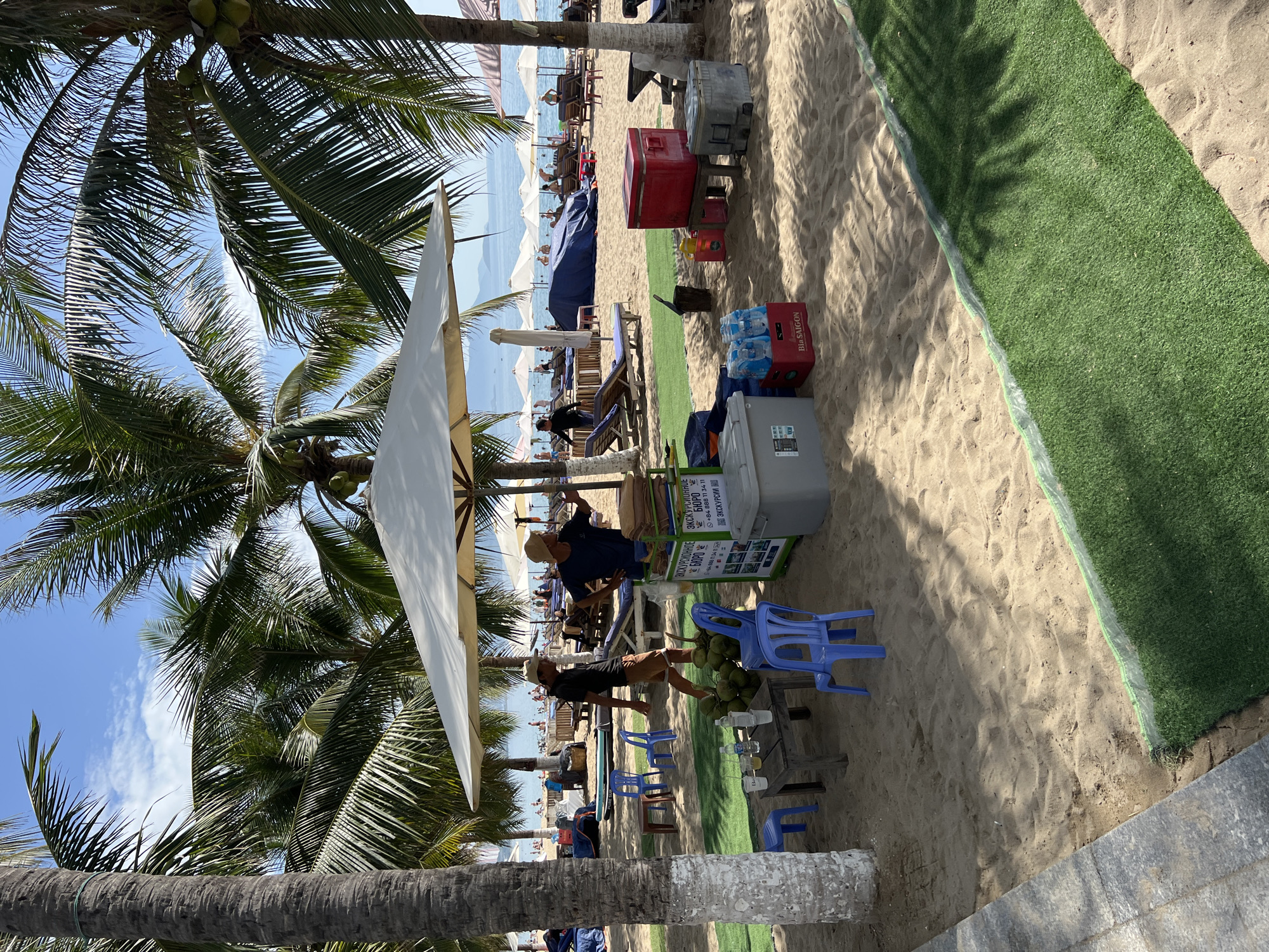 This image captures a sunny day at a beach in Nha Trang, Vietnam. The foreground is dominated by sand and a patch of artificial grass. Several palm trees with coconuts are scattered throughout the scene, casting shadows on the sand.
In the center of the image, there is a large white umbrella providing shade. Beneath it, a small stand displays some information in Russian, possibly advertising tours or services. A few people are visible around the stand, including a man wearing a hat and a dark t-shirt, and another man in a dark t-shirt and a hat.
In front of the stand, there is a table with drinks and cups, and a pile of coconuts. Several blue plastic chairs are placed around the table and stand.
Further back, the beach is lined with lounge chairs and umbrellas, with numerous people enjoying the sun and sea. The ocean is visible in the background, with calm waters and a few distant islands or land masses. The sky is blue with scattered white clouds. There are also several coolers and crates on the sand, some containing bottled water. The overall atmosphere suggests a relaxed beach resort setting.