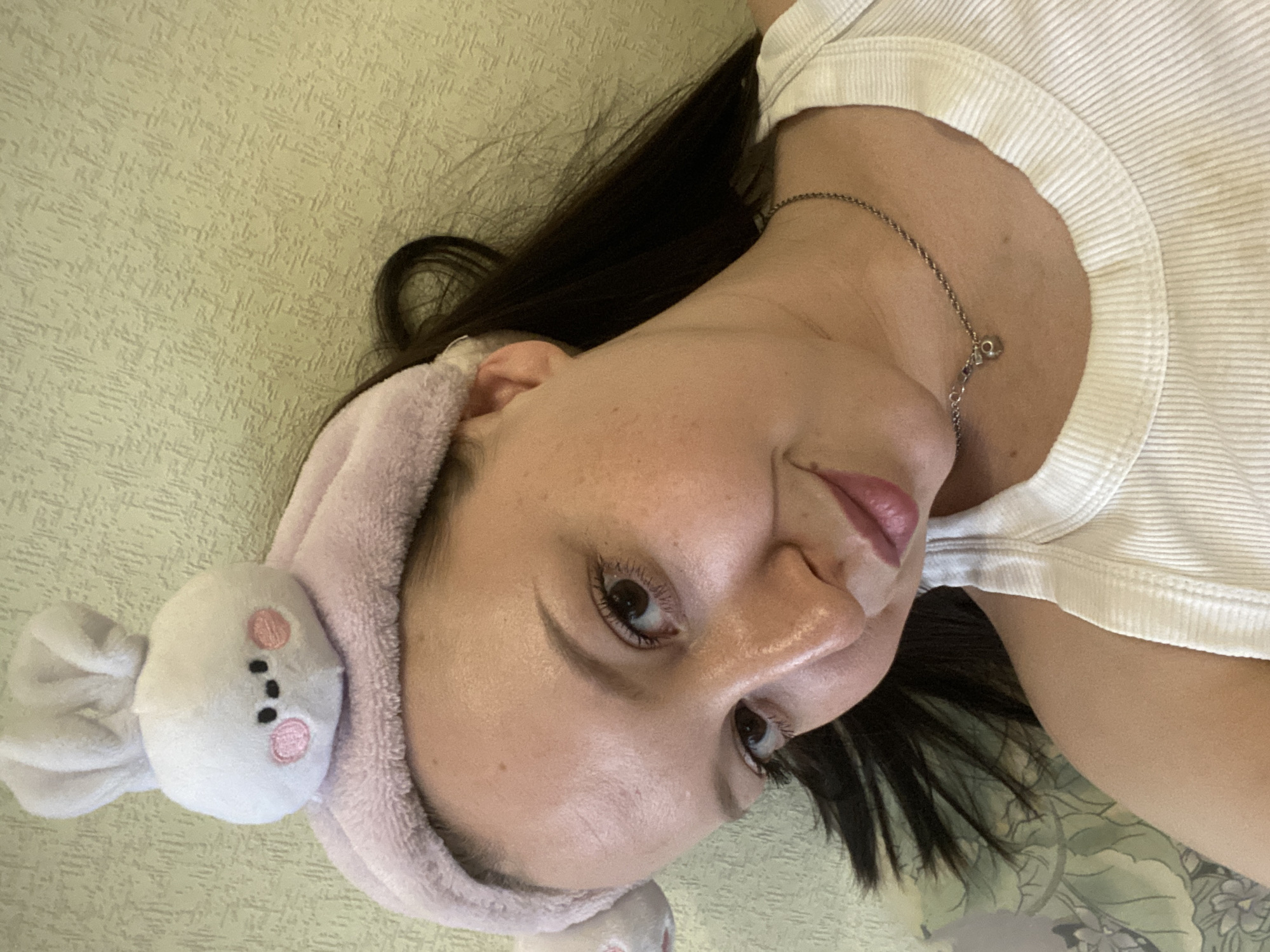 A person is lying down, their head tilted towards the camera. They are wearing a white ribbed tank top and a pink headband with a white plush bunny attached. The person has dark hair and is wearing a silver necklace. The background appears to be a textured green surface, possibly a bedspread or wall. There is no information available to identify the location as Chaikovsky, Russia.
