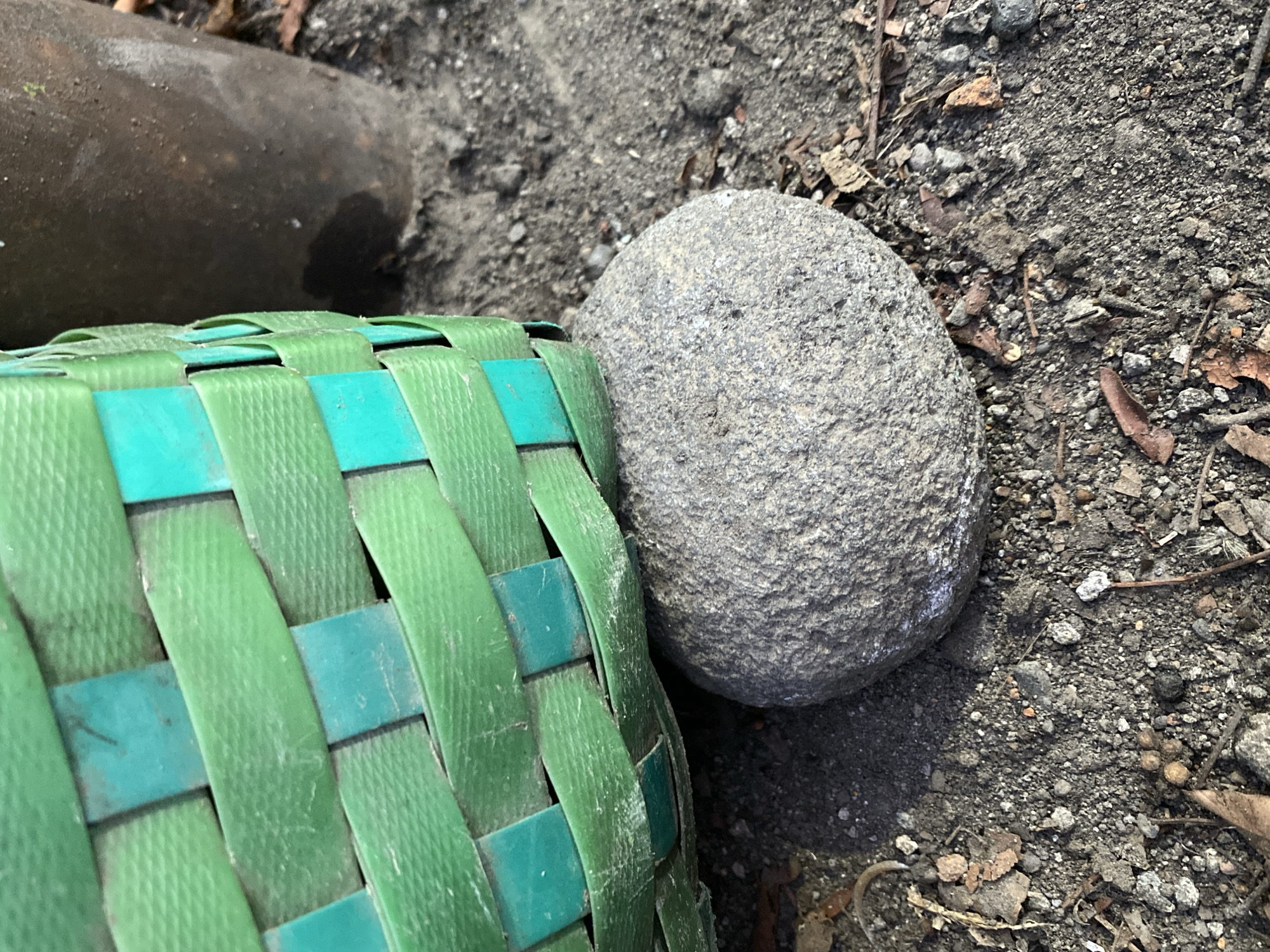 In Kasihan, Indonesia, a close-up shot reveals a textured, grey stone supporting the edge of a green and teal woven basket. The basket, made of plastic strips, shows signs of wear. To the right of the basket, a dark brown, cylindrical object, possibly a pot or container, is partially visible. The ground beneath is a mix of dark soil, small pebbles, and scattered dry leaves and twigs, suggesting an outdoor, possibly garden or yard setting. The lighting indicates a sunny day with shadows cast by the basket and stone. There are no people or animals present in the frame.