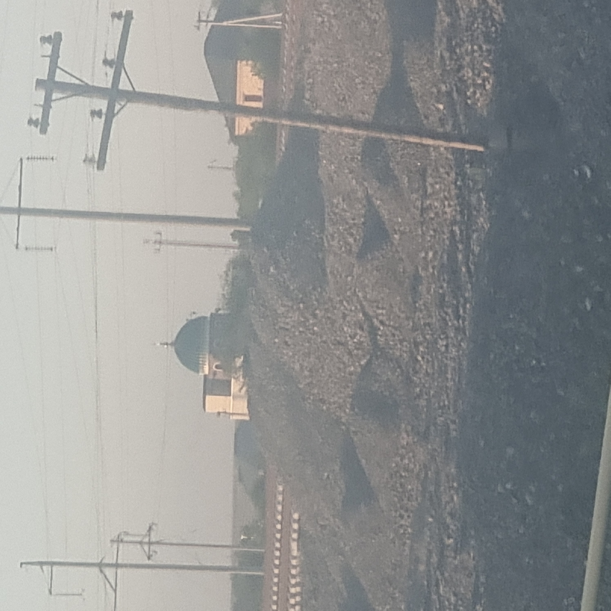 The image depicts a somewhat hazy outdoor scene. The prominent subjects are utility poles with wires crisscrossing the frame, suggesting an infrastructure-heavy environment. In the middle ground, a building with a dome, possibly a mosque or a similar structure, is visible. To the right, a large pile of dark material, likely coal or industrial waste, dominates the landscape. The overall impression is one of a somewhat industrial or semi-rural setting. There are no people visible in the image. The lighting suggests it is daytime, and the haze implies either a dusty atmosphere or overcast weather. Specific location cues are limited, but the provided context places it in Arys, Kazakhstan. There is no visible text in the image.