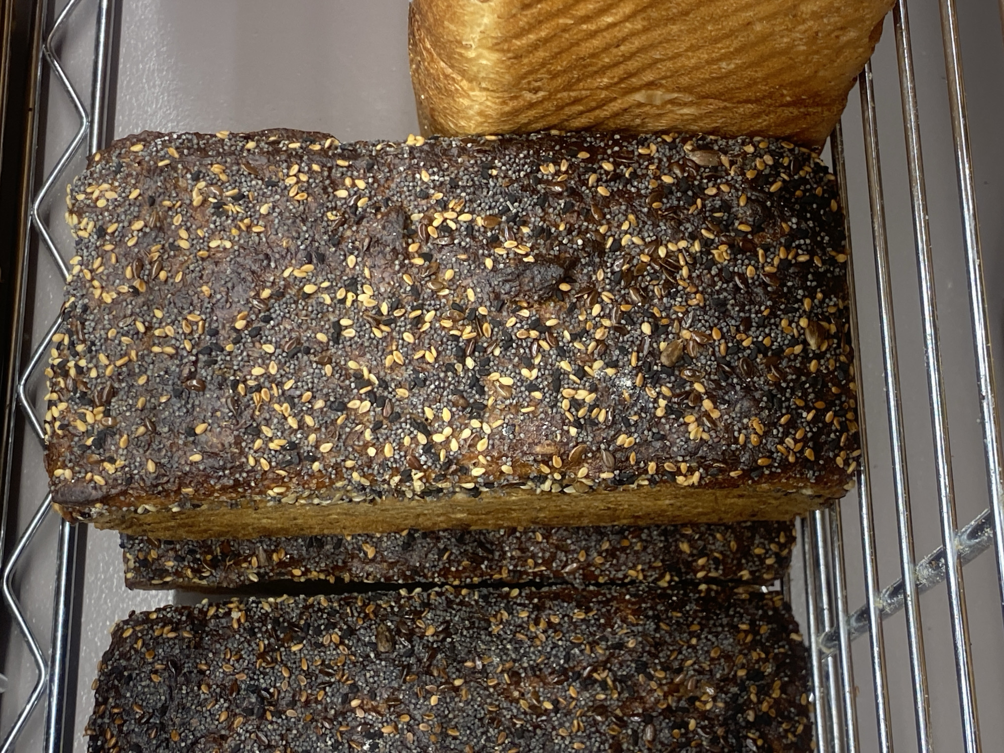 In Warsaw, Poland, on a metal shelf, are three loaves of bread. The two loaves on the left are dark brown and covered in visible poppy seeds and smaller, lighter-colored seeds. The loaf on the far left is partially visible. The loaf in the center is mostly visible, with its crust dark and textured. The loaf on the right is a lighter golden-brown and appears to have a softer crust with visible lines from baking. The loaves are arranged side-by-side.