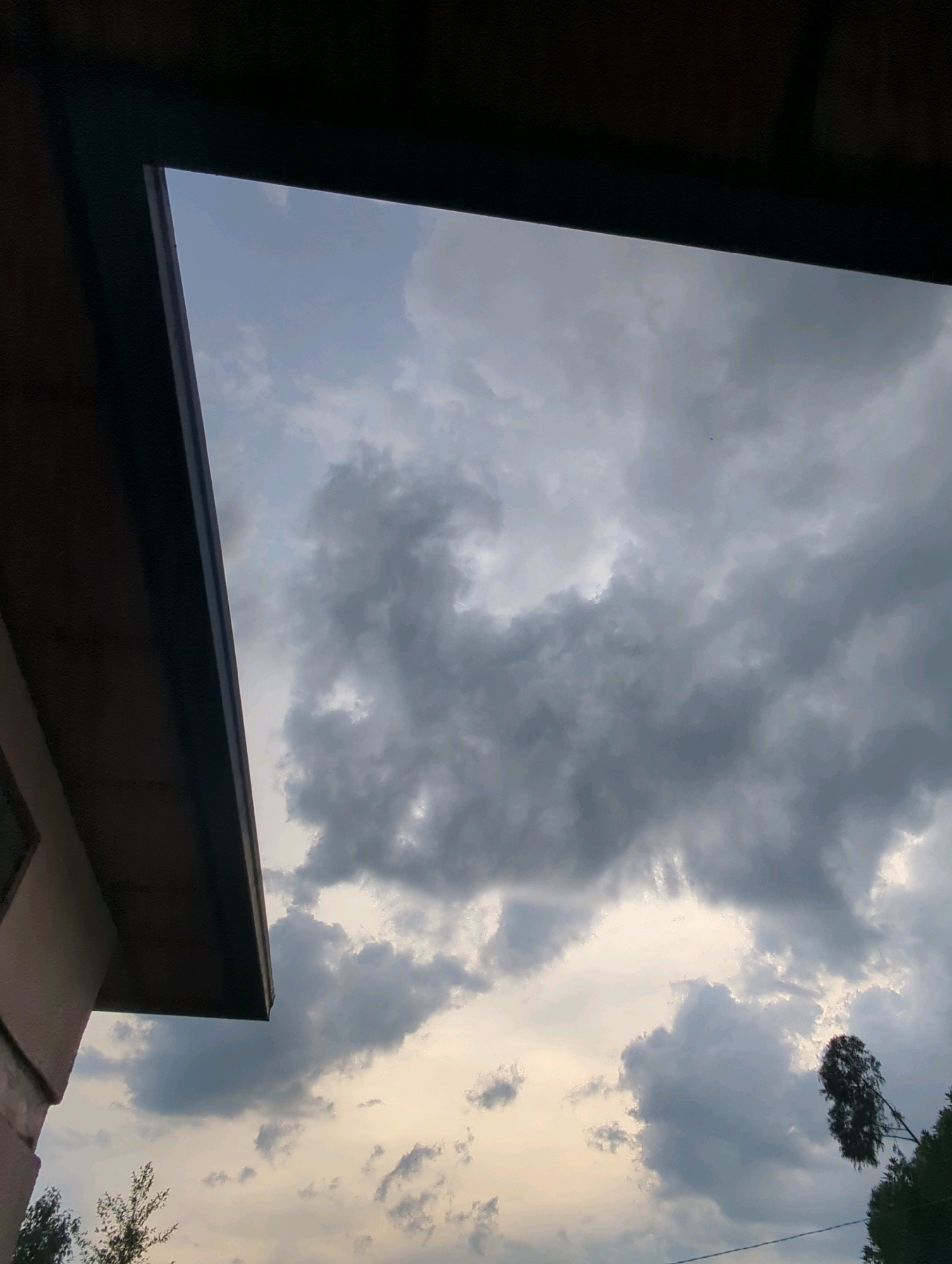 The image captures a dramatic sky filled with heavy, dark clouds, suggesting an impending storm or recent rainfall. A portion of a building's eaves, with a dark brown overhang and a beige wall, occupies the left side of the frame, angling upwards into the sky. The sky itself is a textured canvas of grays and whites, with lighter patches hinting at the sun's presence behind the dense cloud cover. Towards the bottom right, the dark silhouettes of trees, including a distinctive, leaning tree with sparse foliage, are visible against the muted sky. A thin, dark line, possibly a wire, stretches horizontally across the lower part of the scene. The overall impression is one of atmospheric intensity, viewed from a partially sheltered outdoor location in Musanze District, Rwanda.
