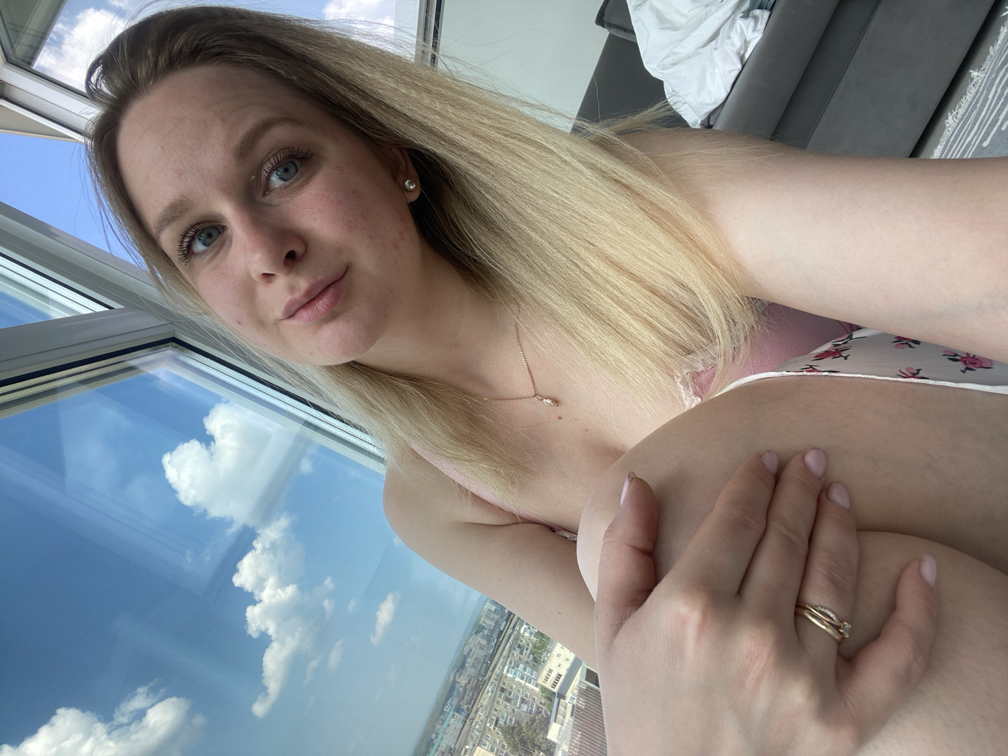 A young woman with blonde hair and blue eyes is pictured in a selfie taken indoors. She is wearing a pink top and a patterned skirt. Her left hand is resting on her leg, showing two gold rings on her finger. Behind her, a large window reveals a cloudy blue sky and a cityscape, which has been identified as Izhevsk, Russia. The room appears to be a bedroom, with a grey sofa and white bedding visible in the background. The lighting suggests it is daytime.
