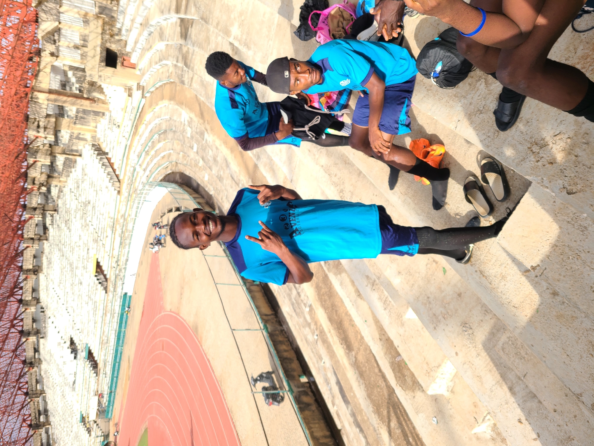 The image shows three young men in blue athletic jerseys standing on concrete stadium steps in Unknown, Unknown. The man in the foreground is smiling and holding up two fingers. The other two men are standing further up the steps, with one appearing to be adjusting his jersey. Various items like a backpack, sandals, and an orange object are near their feet. In the background, there is a running track and a stadium with red overhead structures.