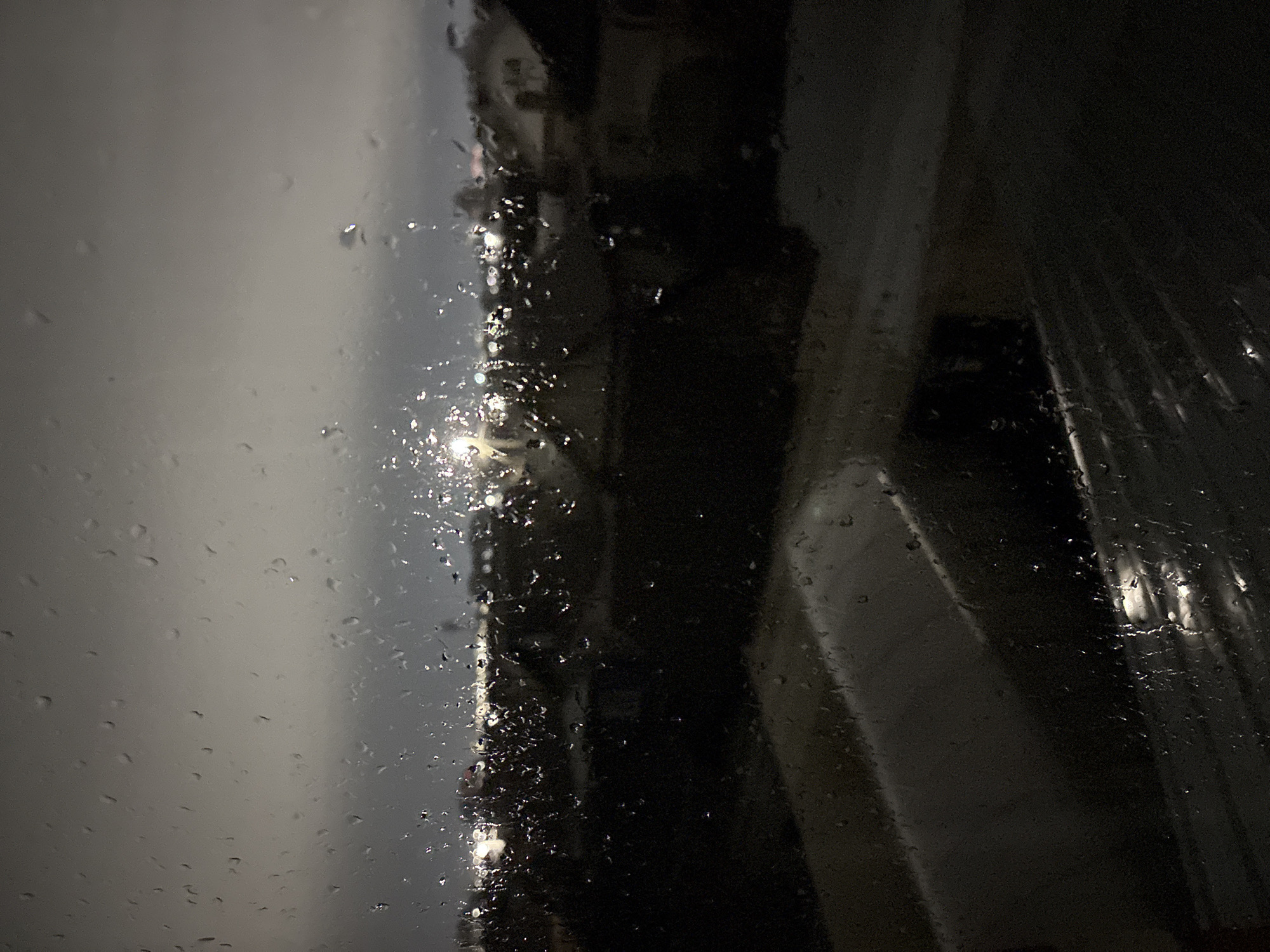 The image shows a rainy night view through a window, with raindrops visible on the glass. Outside the window is a street with lights reflecting off the wet surfaces. Dark buildings are visible in the background. The location is unknown and the presence of the Ulyanovsk city is not possible to be determined from the image. There are no people visible.