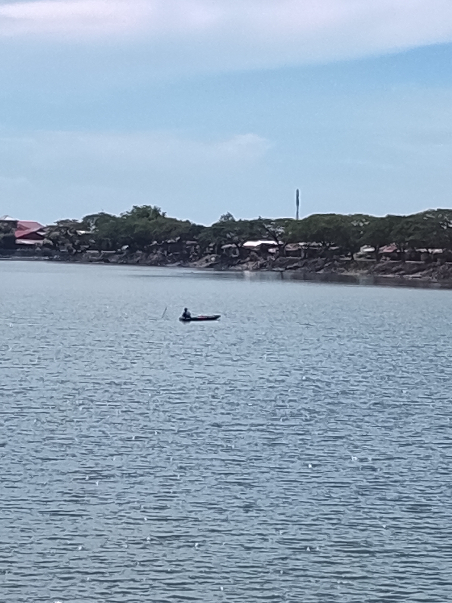 A single person is visible in a small, dark boat on a body of water. The person is silhouetted and appears to be seated. The boat is elongated and appears to be a dugout canoe or similar vessel. The water is calm with some ripple marks. In the background, a treeline with buildings is visible along the shore. The buildings appear to have red roofs and are nestled amongst the trees. A tall, thin structure, possibly a communication tower, rises above the trees in the distance. The sky is a pale blue with some scattered clouds. The scene depicts a tranquil moment on the water with a shoreline settlement in the background. This image may have been taken in Lhokseumawe, Indonesia, a coastal city known for its beaches and water activities.
