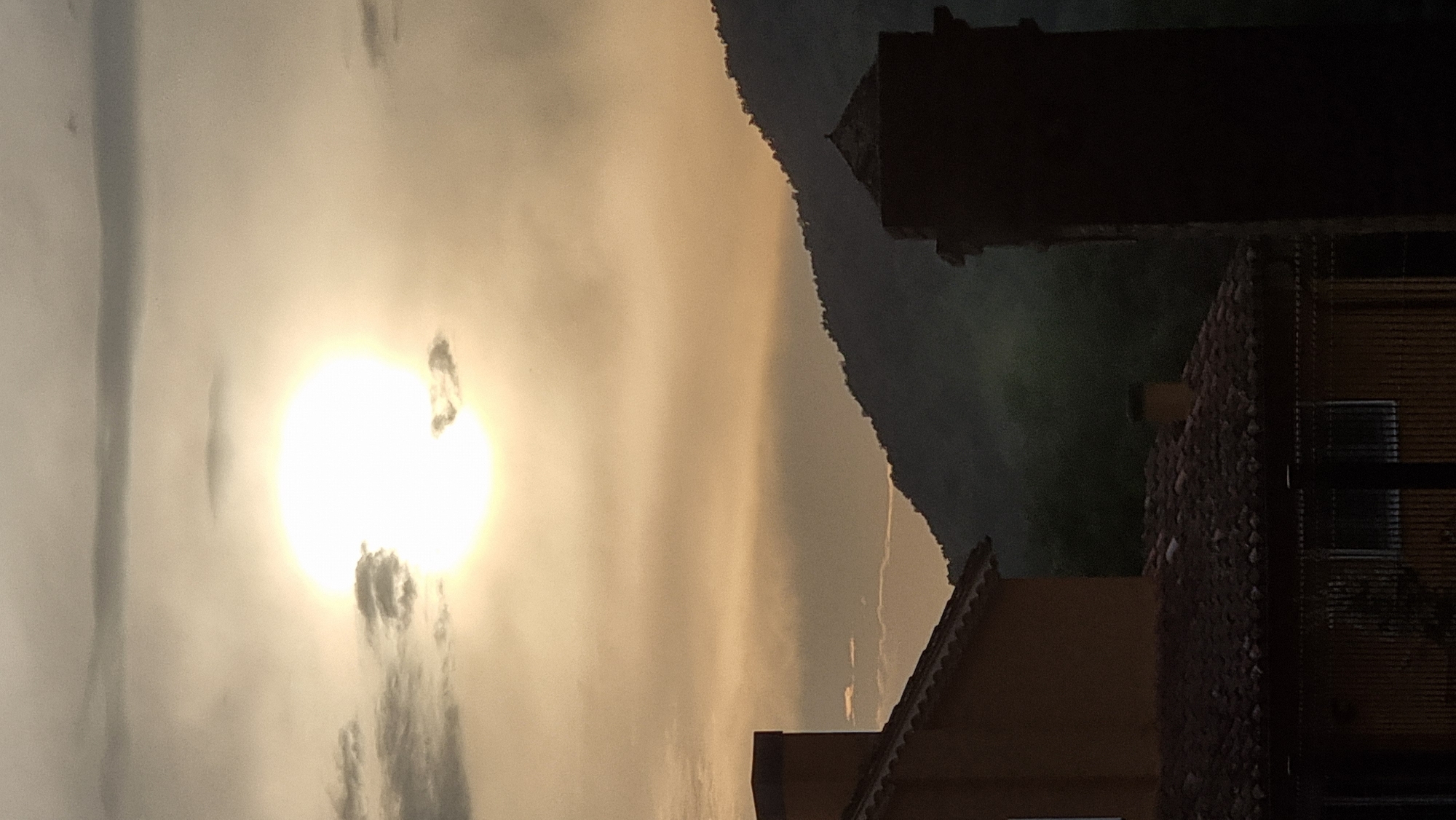 The image depicts a sunset scene with the sun prominently displayed in the sky, partially obscured by clouds. In the foreground, the roof of a building with terracotta tiles is visible, along with part of an orange-colored structure that appears to be a house or part of a complex. To the right, a dark, tall, column-like structure with a cap is silhouetted against the sky. In the background, a dark, densely vegetated mountain range forms the horizon. The lighting suggests it is either sunrise or sunset, with the sun casting a warm, diffused glow. The location is identified as Sant Joan Les Fonts, Spain.