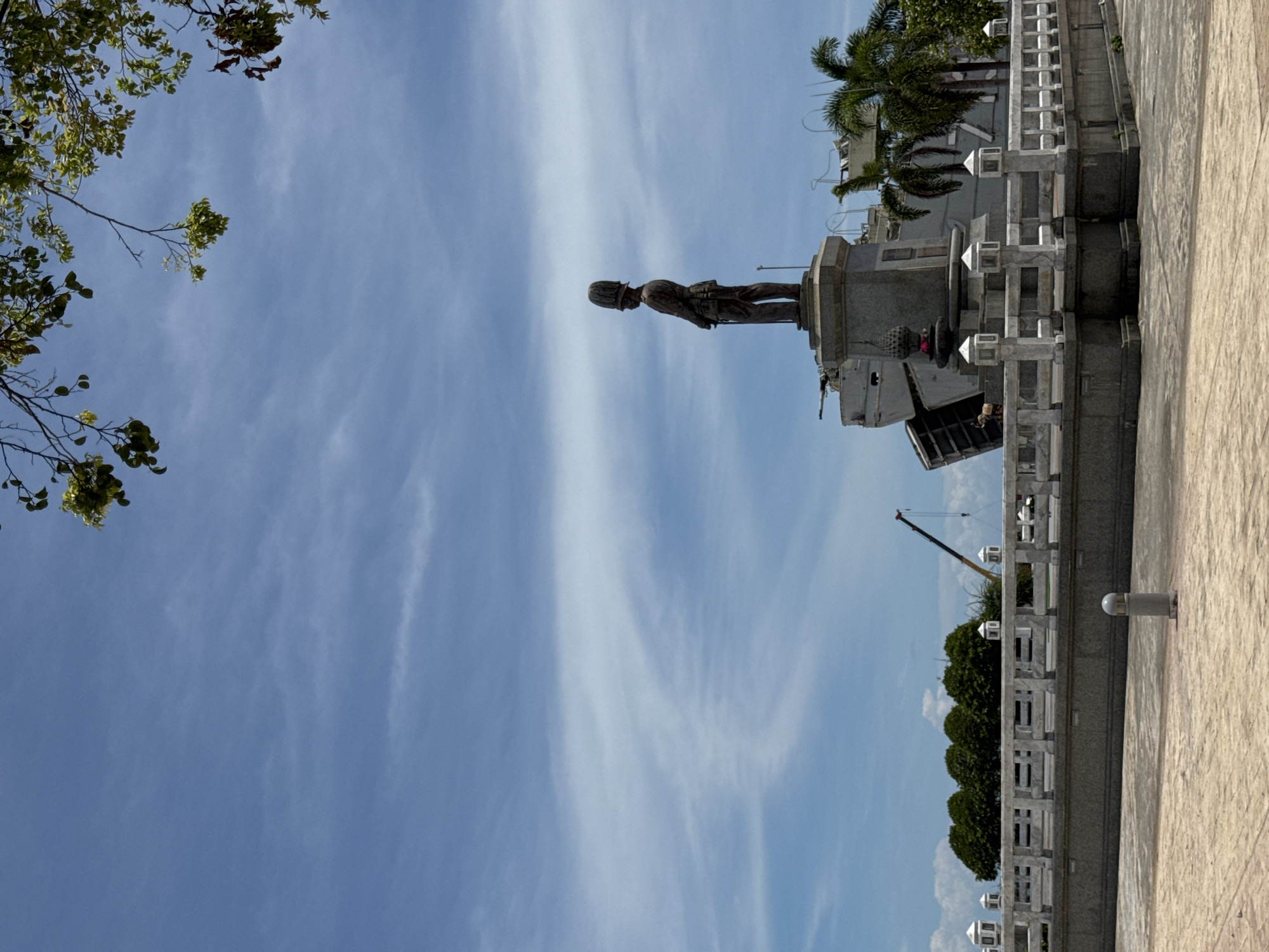 A bronze statue of a man in military uniform stands on a stone pedestal in front of a large grey ship. The statue is facing left in profile. Behind the statue, the ship appears to be docked or grounded, with a crane visible to its left. A white stone railing with lamp posts runs in front of the statue and extends to the right. Trees and palm trees are visible behind the railing. The scene is set on a paved area under a blue sky with wispy white clouds. The image was taken in ตำบลเกาะพะงัน, Thailand.