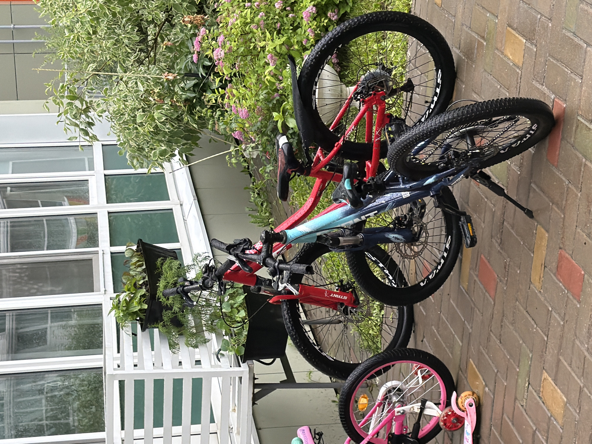 This media file shows two bicycles and a scooter parked next to a building with large windows and a brick pathway. The bicycles are leaning against each other and appear to be mountain bikes. One bicycle is red and blue, while the other is red. The scooter is pink and has decorative elements.
The scene is set outdoors, with lush green foliage and pink flowers visible in the background. The building appears to be modern, with white frames around the windows. The ground is paved with bricks in a herringbone pattern, with some variations in color.
It is difficult to determine the time of day or weather conditions from the image alone, but the lighting suggests it might be daytime. There are no people visible in the image.
Notable details include the brand name "STRIDA" visible on the red bicycle. The overall impression is one of an outdoor recreational area, possibly a backyard or a public space.