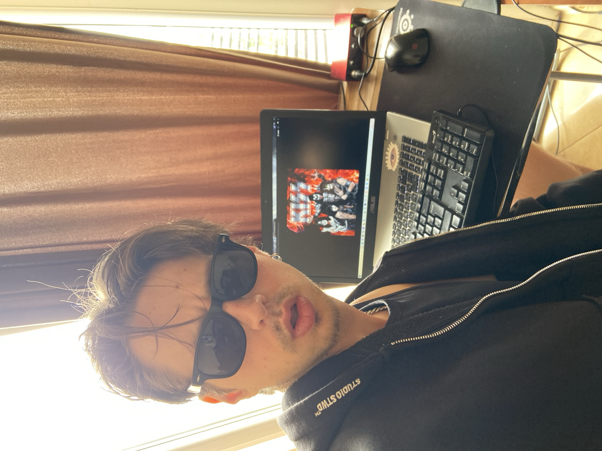 A person wearing sunglasses and a black jacket is positioned in front of a desk. On the desk is a laptop displaying an image of the band KISS, a keyboard, a mouse and a mousepad. A red object is also visible near the window. A brown curtain is in the background. The location is not discernible from the image.