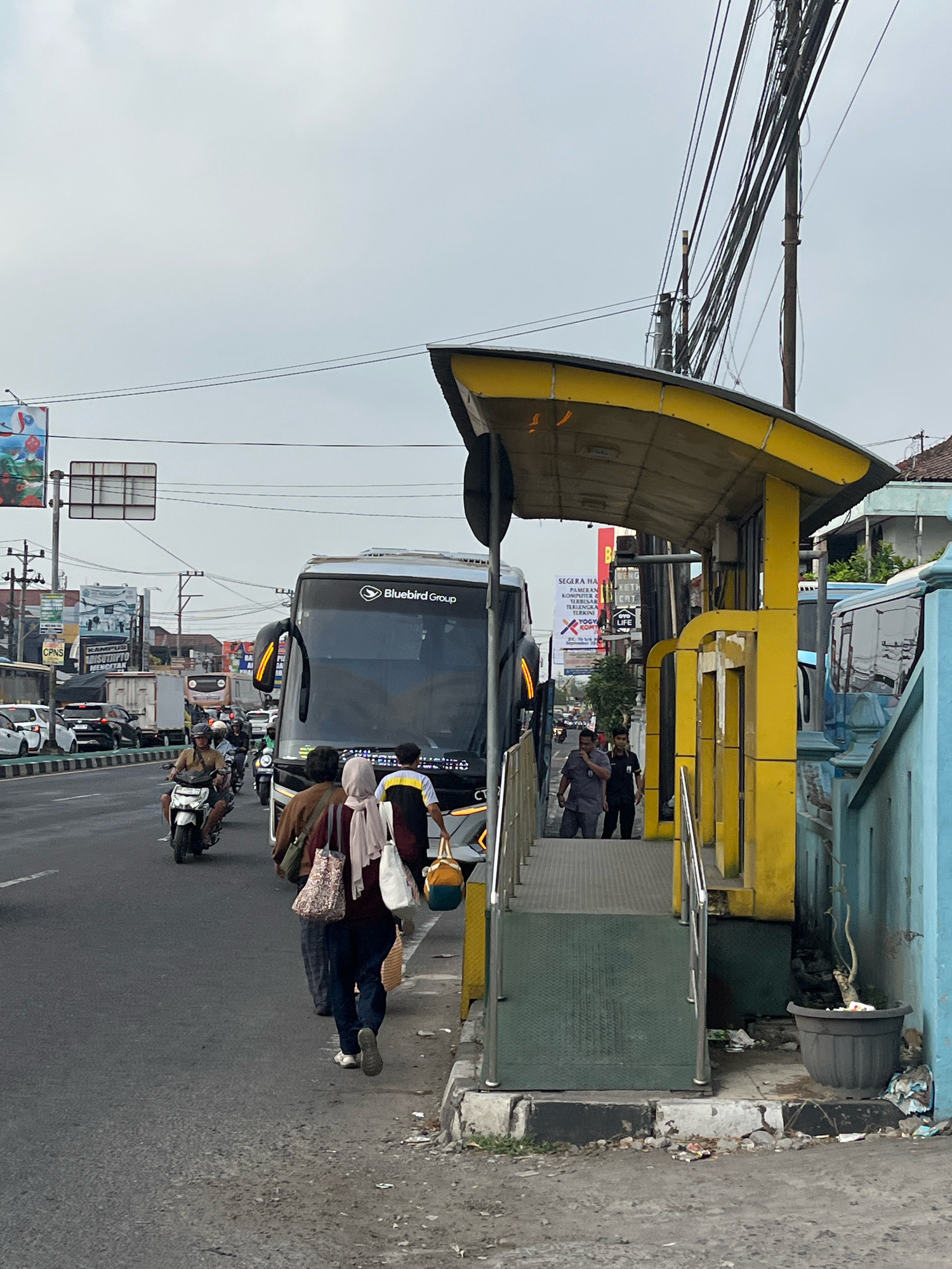 A street scene in Banguntapan, Indonesia, depicts a bus stop with a yellow awning. A large blue and silver bus, identified as "Bluebird Group," is stopped at the bus stop, partially blocking the view of other traffic. Several people are visible: two individuals are walking away from the bus stop on the sidewalk, one carrying a patterned bag and the other a white bag. Two men in dark clothing are standing near the entrance of the bus stop. In the street, a person is riding a white motorcycle and another is on a scooter. Other vehicles, including cars and a truck, are visible in the background, indicating moderate traffic. The environment appears urban, with power lines overhead and buildings along the side of the road. Trash is scattered on the sidewalk and street. The sky is overcast.