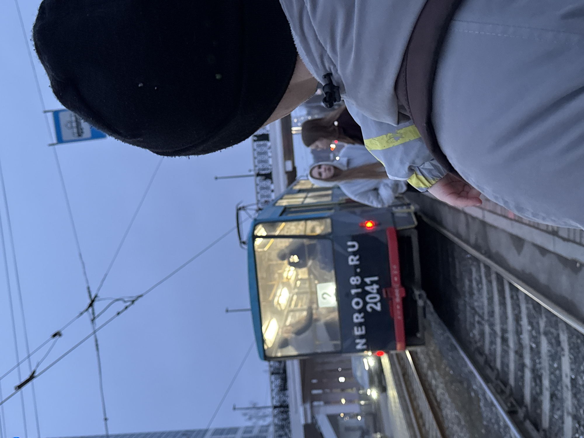 A northward-oriented view shows a light rail train stopped on tracks in Izhevsk, Russia. The train is numbered 2041 and marked with the website "NERO18.RU". Overhead wires and a blue sign featuring a bus icon are visible. Two girls stand near the train. A person wearing a black cap and gray jacket partially obscures the view.