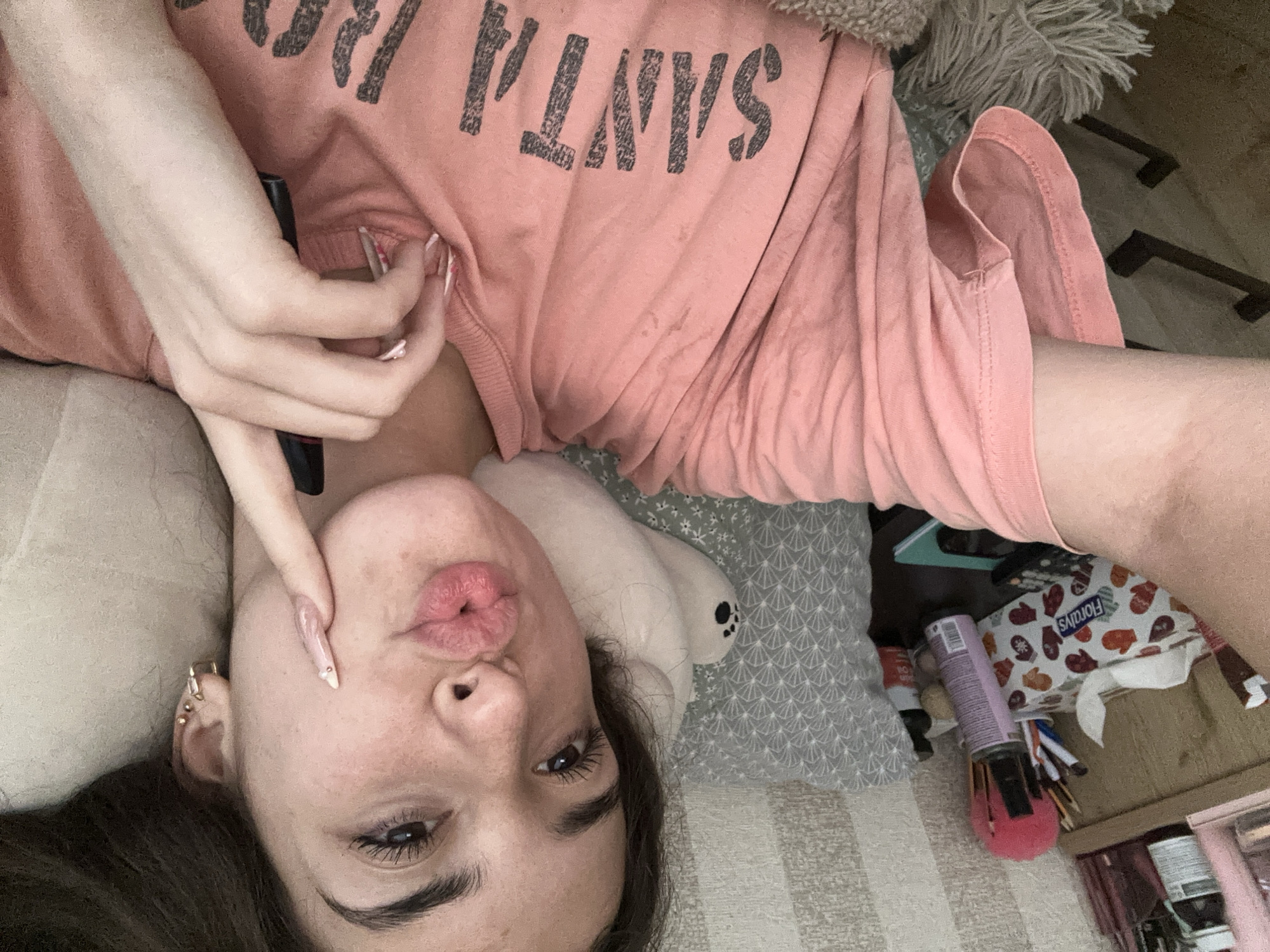 A young woman with dark hair is lying down, taking a selfie. She is wearing a pink t-shirt with the word "SANTA" visible on it. Her left arm is extended, holding the camera. Her right hand is raised to her cheek, with a finger touching it. She is making a kissing face. The background shows a bedside table with a tissue box, makeup, and a remote control. A striped wall and a pillow are also visible. The scene is indoors, possibly a bedroom. There is no indication of the location in the city of Shumen, Bulgaria.
