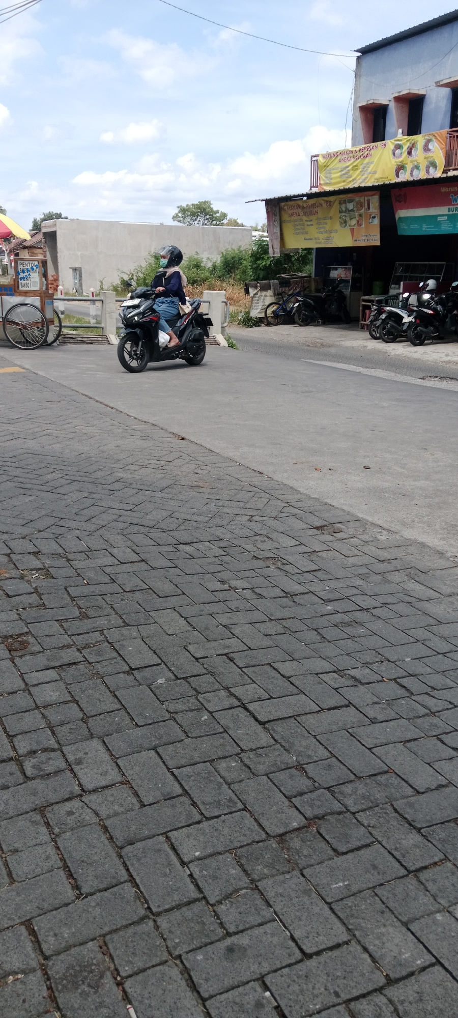 The image depicts a street scene in Maguwoharjo, Indonesia, under a cloudy sky. In the foreground, a paved road with a distinctive herringbone pattern of dark grey bricks is visible. A person wearing a blue long-sleeved shirt, blue jeans, a face mask, and a black helmet is riding a black motorcycle. They are positioned in the middle of the road.
To the left of the road, a white cart with a spoked wheel is parked. Behind the cart, a low concrete wall runs parallel to the road. Further in the background, a row of parked motorcycles is visible in front of a building with a blue and orange facade. A yellow banner with text and images is displayed on the building, suggesting a food establishment. The building has multiple windows with orange frames. Beyond the building, trees and vegetation can be seen. The overall impression is a typical urban street in Indonesia, with everyday elements of transportation and commerce.