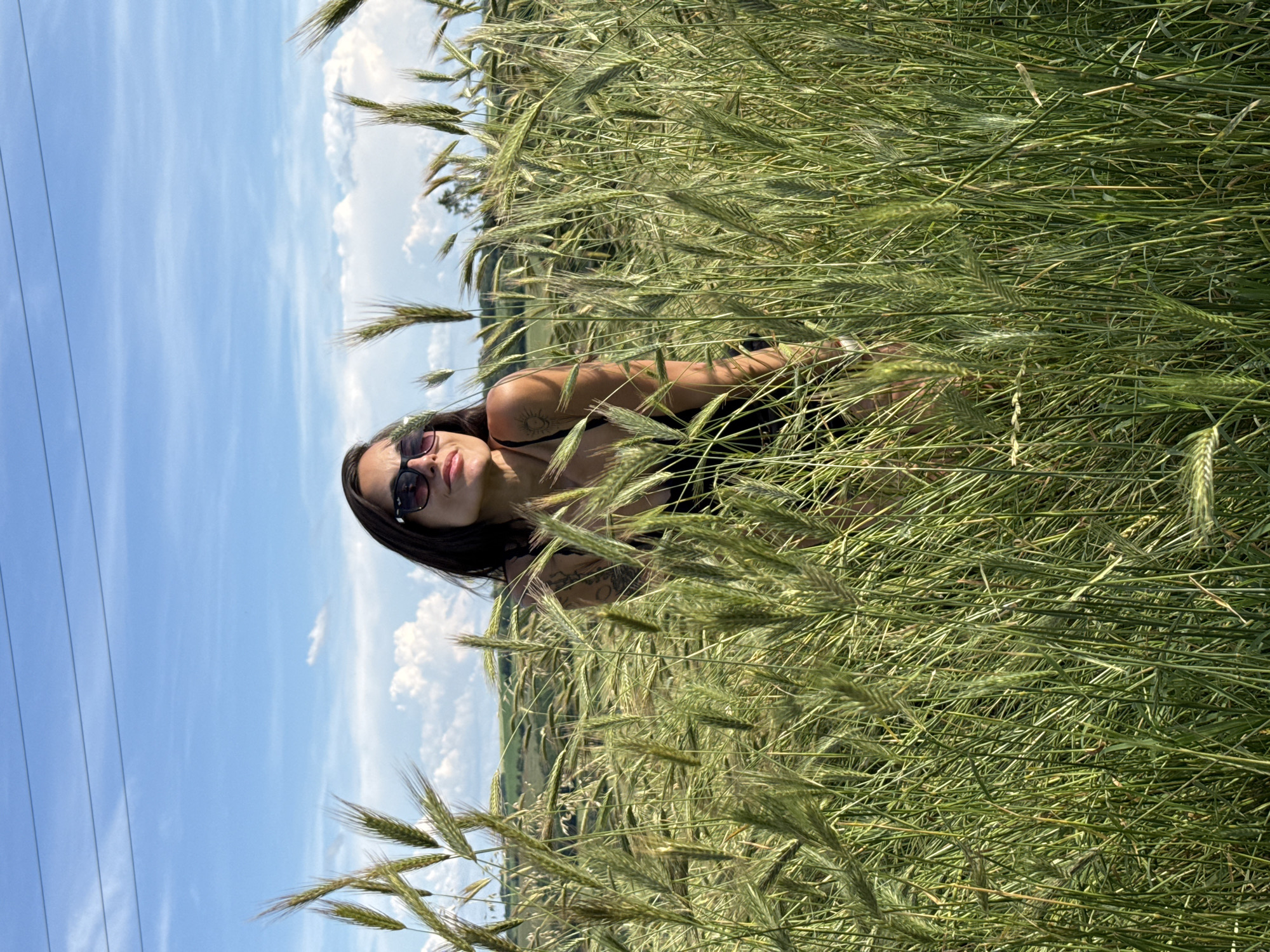 A young woman with dark hair and sunglasses is lying down in a field of tall, green grass. She is wearing a black bikini top. Her body is partially obscured by the dense foliage, but her face and upper torso are visible. She is looking towards the camera with a relaxed expression. In the background, the sky is a clear blue with scattered white clouds. There are power lines visible in the upper left portion of the image. The overall scene suggests a warm, sunny day in a rural setting, likely during the summer. The image appears to be taken in Pleven, Bulgaria. There is no visible text in the image.