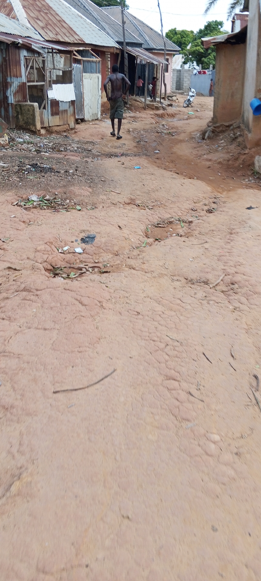 The image depicts a dirt road in Keffi, Nigeria, lined with rudimentary structures built from corrugated metal and brick. An adult male with a bare torso and dark clothing walks away from the camera. Several children stand in the doorways of the buildings in the background. Trash litters the foreground and the roadside. The road surface is uneven and appears to be made of packed earth.