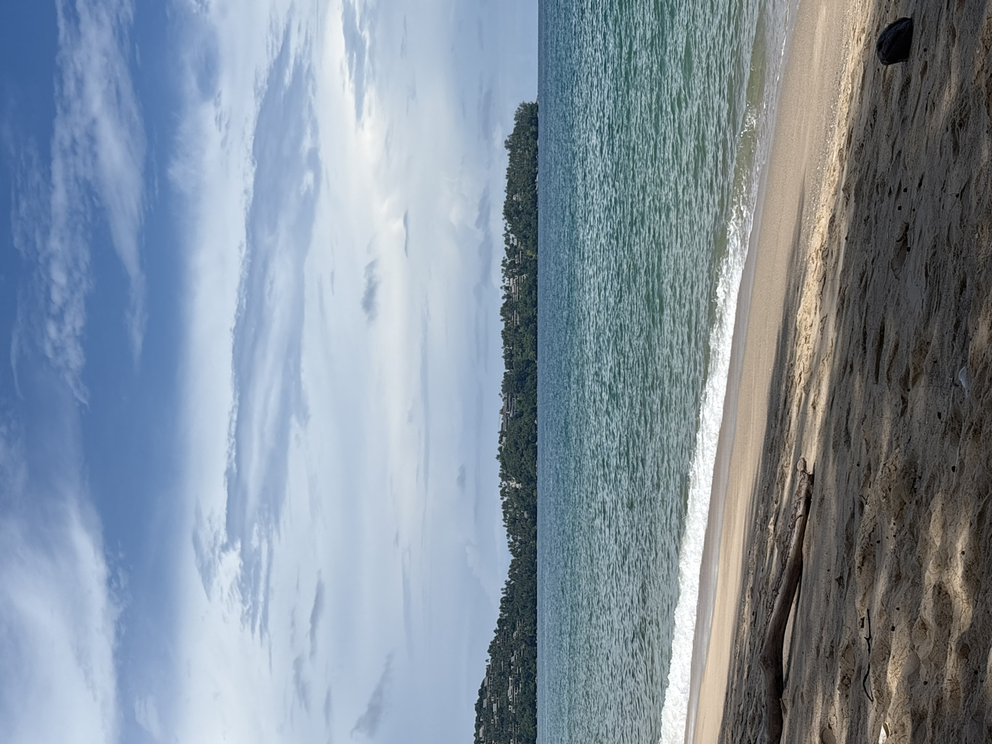 A wide-angle, full-shot photograph captures a serene coastal scene in Choeng Thale, Thailand. The image is divided into three horizontal bands: sky, sea, and beach. The upper portion of the frame features a blue sky with scattered, wispy white clouds, some appearing more dense than others. Below the sky, a tranquil turquoise sea stretches across the mid-ground, with gentle waves lapping at the shore. The water exhibits a slightly rippled texture, suggesting a light breeze. Along the distant horizon, a densely wooded headland is visible, dotted with numerous buildings that appear to be resorts or residential structures. The foreground is dominated by a sandy beach, rendered in shades of light brown and beige. The sand shows some undulations and is partially shaded, casting dappled shadows across its surface. A single piece of driftwood, elongated and weathered, lies diagonally across the left side of the beach. Towards the right foreground, a dark, rounded object, possibly a coconut, rests on the sand. The scene is devoid of any visible people or animals. The overall impression is one of natural beauty and peaceful seclusion.