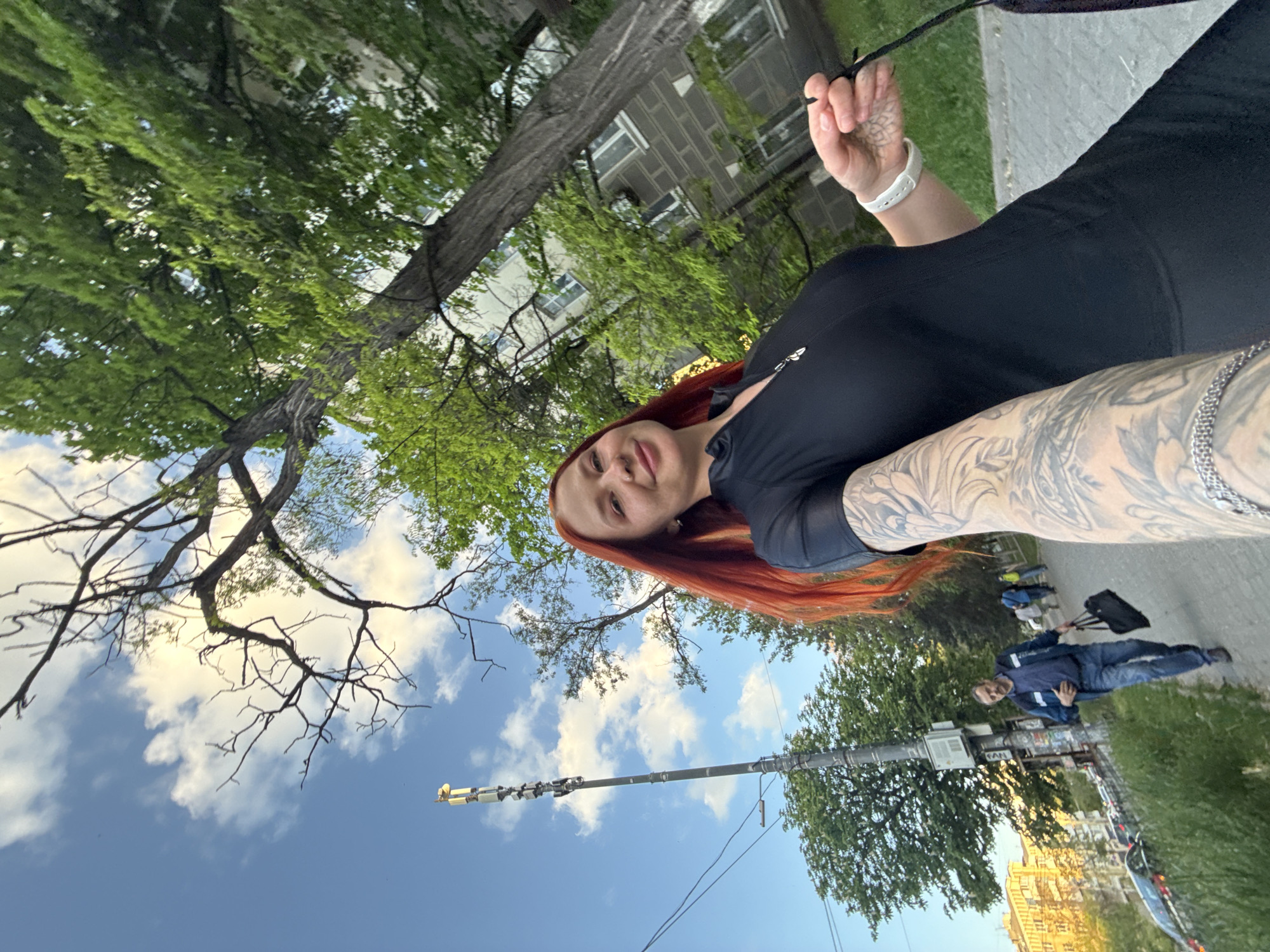 A rotated, medium-shot selfie shows a red-haired woman with extensive arm tattoos holding a black bag, standing on a sidewalk in Rostov-on-Don, Russia. The sky is visible with scattered clouds and partially obscured by tree branches. In the background, a man is walking on the sidewalk with a bag. A building is also visible in the distance. A utility pole is standing on the side of the road.