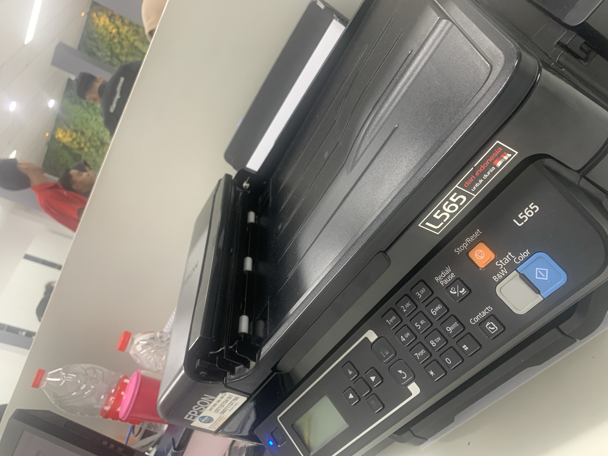 The image shows an Epson L565 printer on a desk in what appears to be an office environment. The printer is black and has various buttons and a small LCD screen on its control panel. A sticker with "L565 dari indonesia untuk dunia" (L565 from Indonesia for the world) is visible on the side of the printer. In the background, there are people, water bottles, and office furniture. The context of "Palembang, Indonesia" is not directly evident from the image itself but is provided as an external piece of information.