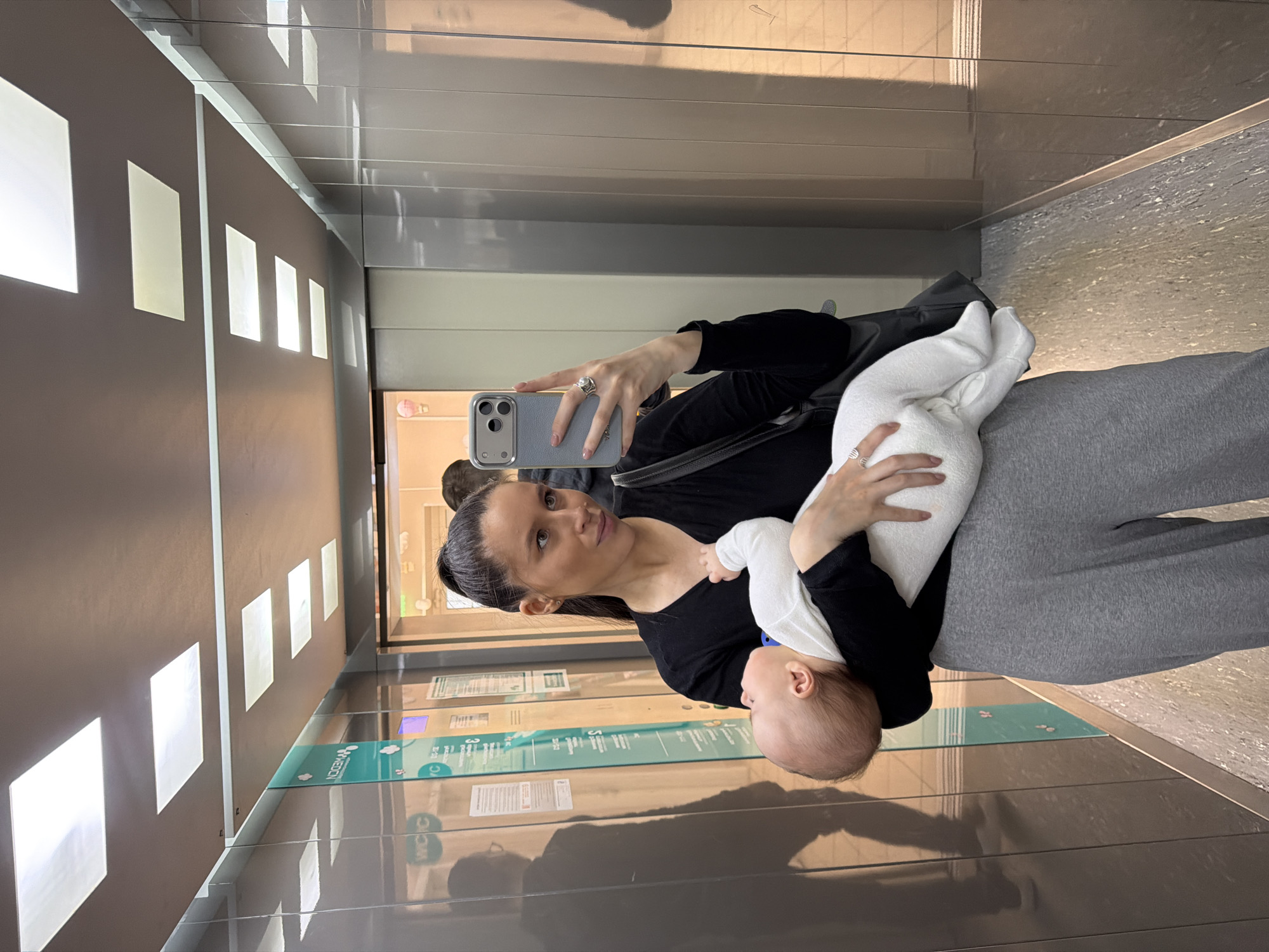 The image shows a woman and a baby inside an elevator. The woman is holding the baby in one arm and taking a selfie with a phone in her other hand. The elevator has reflective metal walls and recessed lighting. Based on the information visible through the elevator doors, the location appears to be Moscow, Russia, possibly an airport due to the signage.