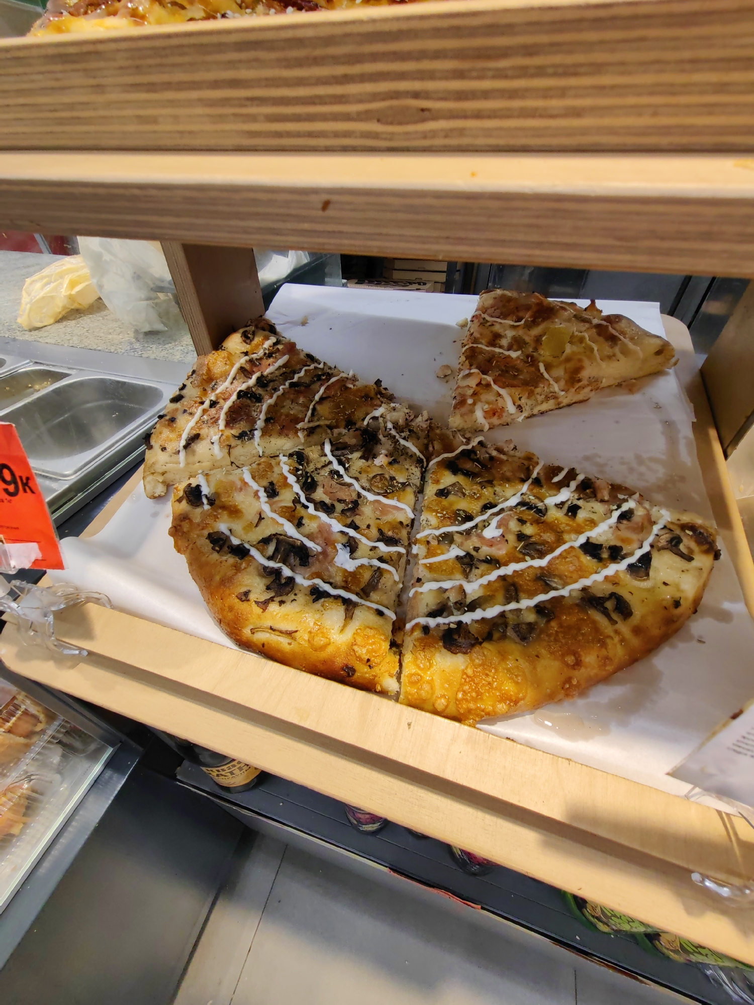The image shows a close-up view of several slices of pizza on a white paper liner on a wooden shelf. The pizza appears to be a large, round pie that has been cut into slices. The toppings visible include mushrooms, what looks like bacon or ham, and white sauce drizzled over the top in a zigzag pattern. The crust is golden brown and appears to be slightly crispy. In the background, there are stainless steel containers and a red tag with white text that is partially visible. The overall setting seems to be a food display, possibly in a grocery store or bakery. The lighting is bright, suggesting an indoor environment. There are no people visible in the frame. The focus is entirely on the food.