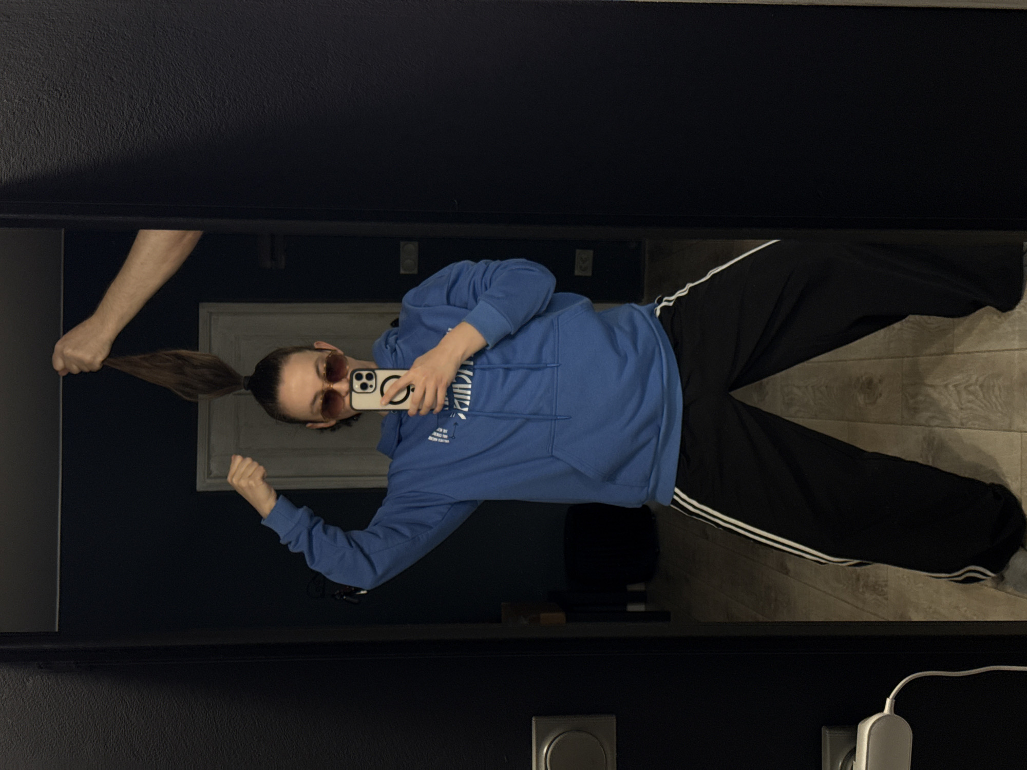The image, originally rotated 90 degrees counter-clockwise, captures a mirror selfie taken indoors in Chaikovsky, Russia. The primary subject is a person, likely female, standing in a casual pose. She is wearing a light blue hooded sweatshirt with faint, unreadable white text across the chest, and black athletic pants with two white stripes down the side. She sports sunglasses with reddish-brown tinted lenses and has her hair pulled back into a ponytail. She holds a smartphone, likely an iPhone with a distinctive camera array and a textured case, to capture the reflection.
A second person's bare arm is visible on the left, playfully holding the primary subject's ponytail, adding a lighthearted and interactive element to the scene.
The background visible in the mirror's reflection includes dark-colored walls and a light wooden floor. A white doorframe is partially seen behind the subject. In the foreground of the reflection, a grey circular object, possibly a smart home device or thermostat, and a white charging cable connected to a wall outlet are visible. The lighting is dim, suggesting an indoor setting without strong natural light. The overall mood is relaxed and informal.