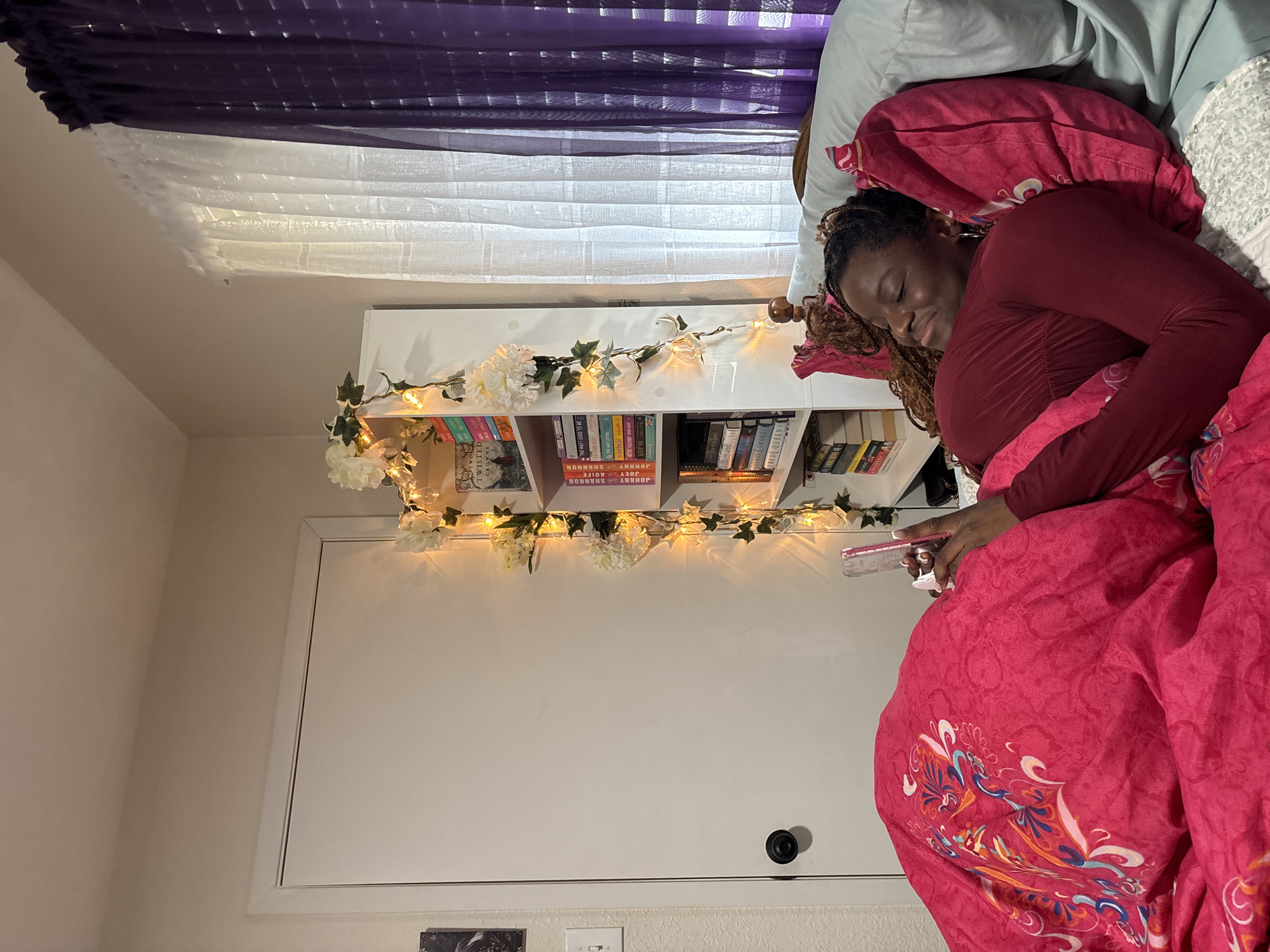 This image captures a serene bedroom scene, likely located in Arlington, United States of America, showing an adult individual sleeping peacefully. The subject, an individual with dark skin and braided hair, is depicted lying on their right side with eyes closed, indicating deep sleep. They are wearing a maroon long-sleeved top and are covered by a bright pink duvet featuring an intricate, colorful pattern, possibly a peacock design. Their head rests on a matching pink patterned pillow, with a light blue pillow visible behind it.
To the left of the bed, a white horizontal bookshelf serves as a headboard, decorated with a string of warm fairy lights and artificial white flowers intertwined with green ivy. The shelves are filled with a collection of books. Legible titles include "TWISTED CROWNS," "THE LAST LIFE," "THE GOOD GIRL'S GUIDE TO MURDER," "VERITY," "THE FOURTH WING," and "REST QUEEN." Several books by "JOHNNY SHANNON" are also visible.
Above the bookshelf, a window is dressed with sheer white blinds and dark purple sheer curtains, allowing diffused natural light to filter into the room. A white door with a black doorknob is partially visible below the bookshelf. The sleeping person holds a smartphone with a clear case in their right hand, suggesting recent use. The overall scene conveys a calm and cozy atmosphere, with the person appearing relaxed and at peace. The presence of natural light suggests it is daytime, though the person is asleep.