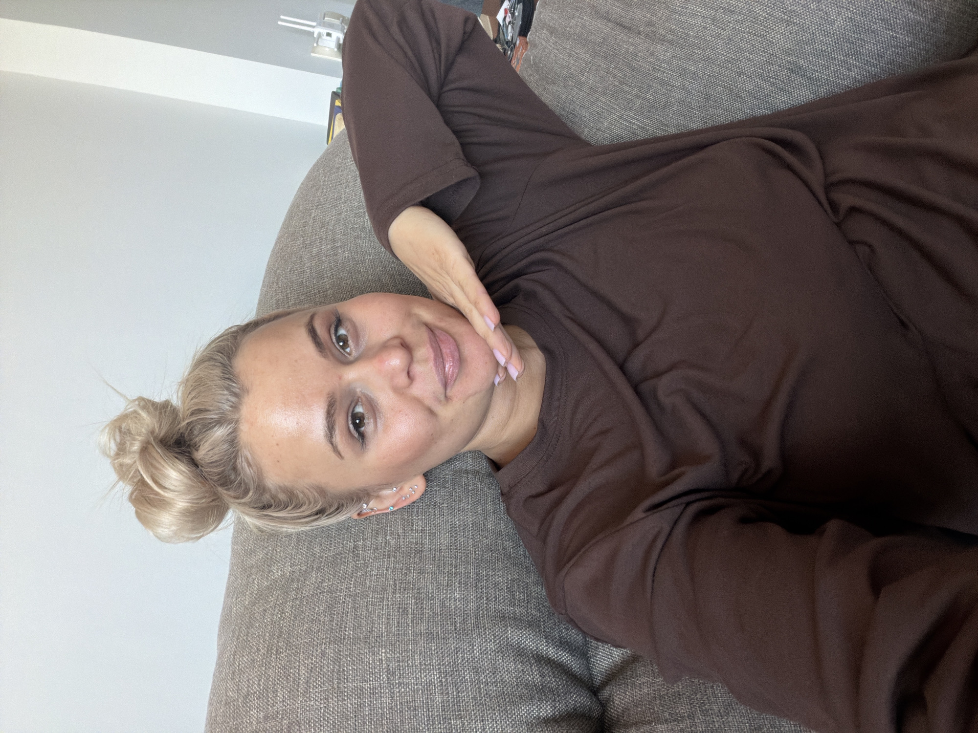 This media file is a selfie of a woman lying on a textured, light gray sofa in an indoor setting in Sarapul, Russia. The woman, who has blonde hair pulled back into a messy bun, fair skin, and light-colored eyes, is wearing a plain brown long-sleeved top. Her left hand, adorned with light pink nail polish, is gently resting near her chin, framing a soft, pleasant smile. She appears relaxed and content. Multiple small, sparkling piercings are visible on her left ear. The background consists of a bright white wall, with a ceiling light fixture partially visible in the top right corner, indicating a well-lit space. The lighting suggests it is daytime. There is no visible text in the image.