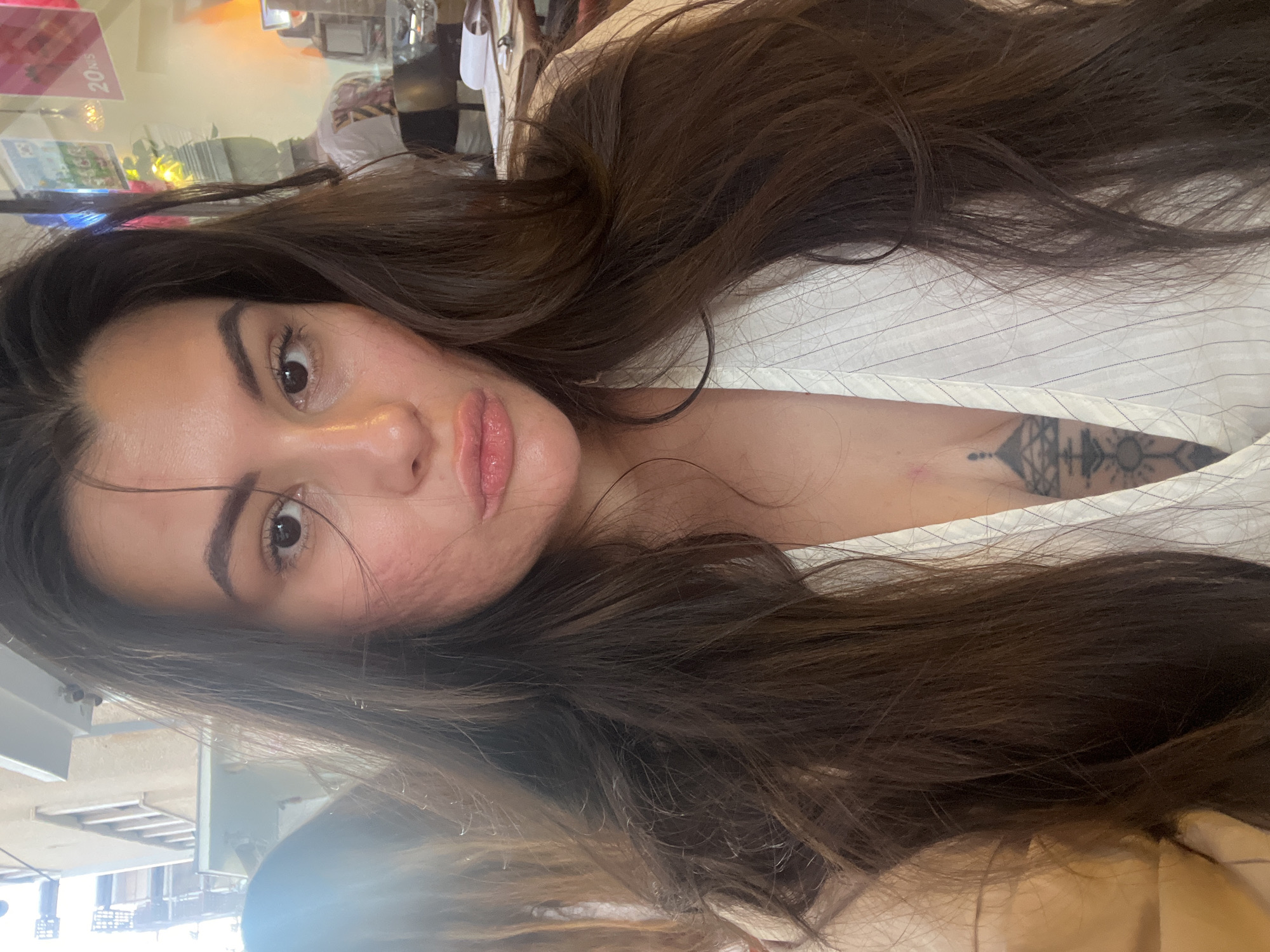 A woman with brown hair is pictured in an indoor setting that appears to be in Tel-Aviv, Israel. She is wearing a white striped shirt. A tattoo is visible on her chest. The background shows a blurred scene suggestive of a retail environment.