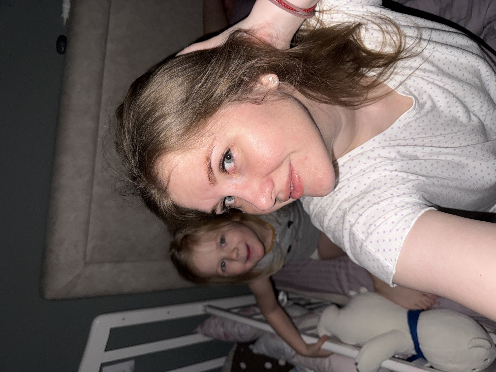A selfie shows a woman and a young child in a bedroom. The woman has light brown hair, fair skin, and is wearing a white shirt with light purple polka dots. She is smiling slightly at the camera. Behind her and below is a young child with blonde hair and a gray shirt, also smiling and looking at the camera. They appear to be on a bed with a white frame. A stuffed polar bear with a blue ribbon around its neck lies next to the child. The bedroom's background includes a gray headboard and what appears to be a wall. The location in the city of Gornyi (Dvurechenskoe S/P), Russia, cannot be determined based on the image.