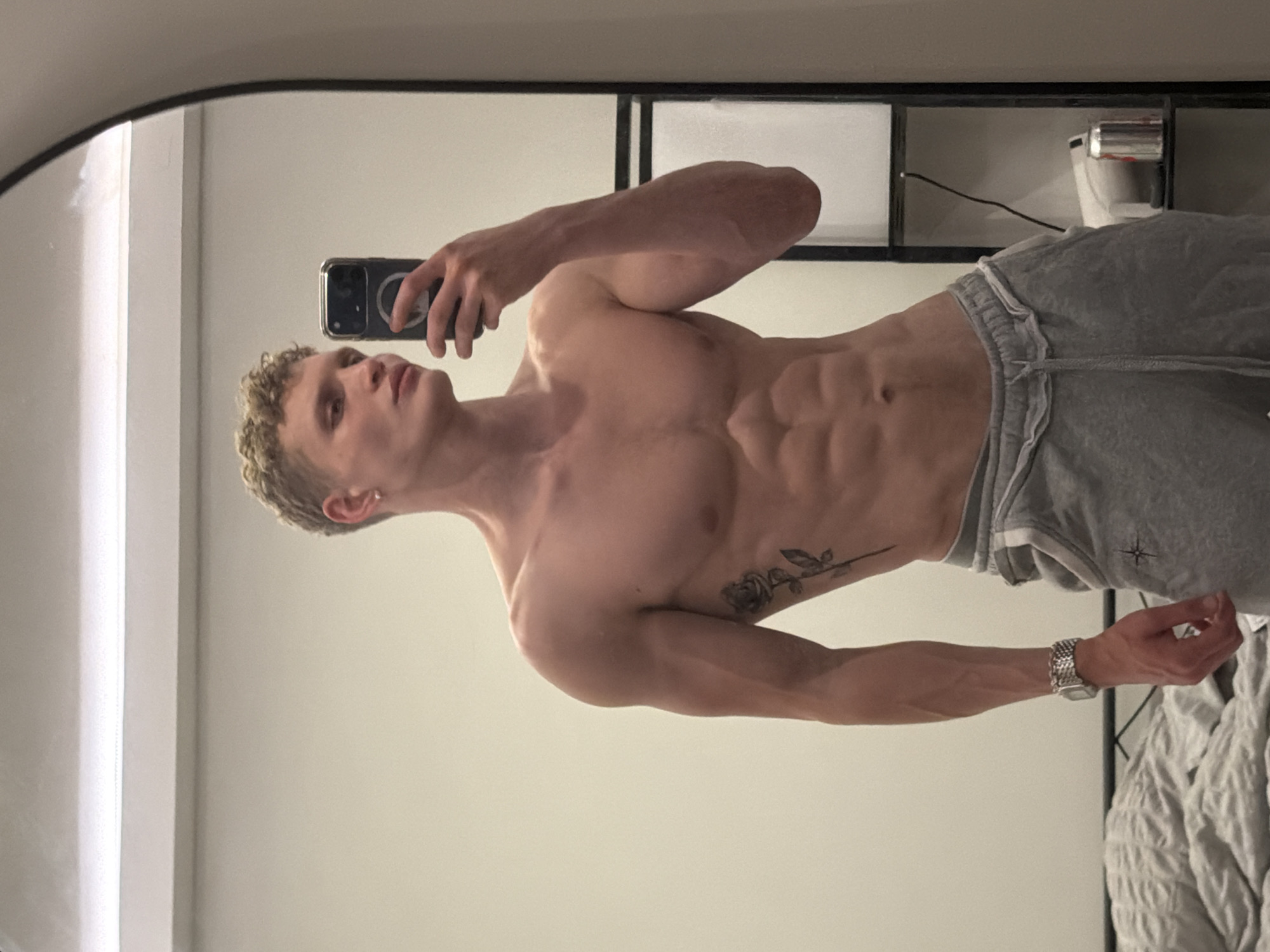 A Caucasian male with blonde, curly hair is standing indoors, reflected in a mirror. He is taking a selfie with his phone held in his right hand. The man is wearing gray sweatpants but is shirtless, revealing a well-defined physique with visible abdominal muscles and a tattoo of a flower on his left side. He is also wearing a silver watch on his left wrist. The background shows a simple white wall and a partially visible black shelving unit with various items on it, including what appears to be a can. The location is indiscernible and I cannot provide more information about London, United Kingdom.