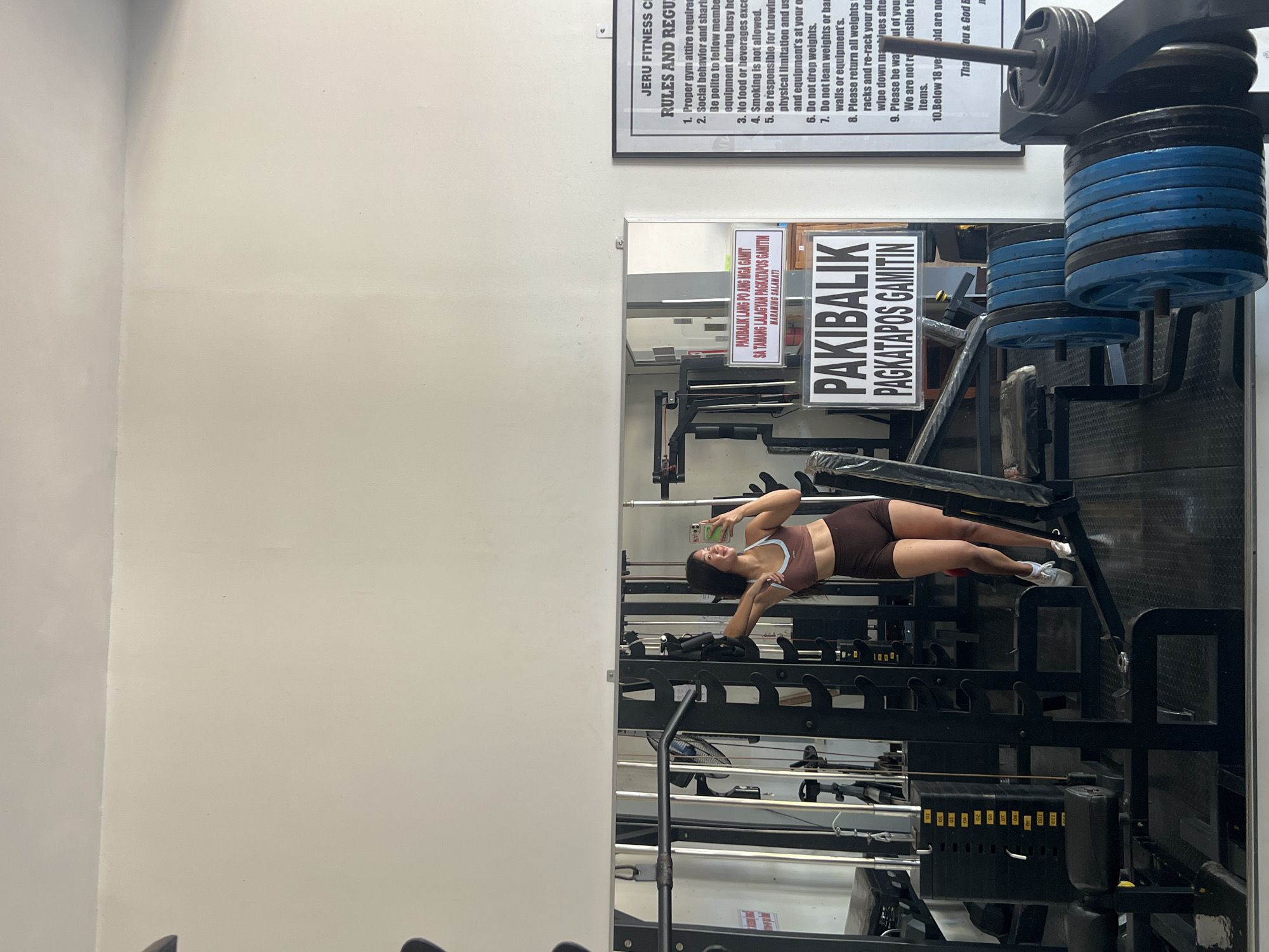 The image depicts a young woman in athletic wear taking a mirror selfie in a gym. She is wearing a brown sports bra and matching shorts, with white athletic shoes. She is leaning against a weight rack, with a smartphone in her right hand. The gym appears to be well-equipped with various weight machines and free weights, including stacks of weight plates in blue and black. A large mirror reflects the gym equipment and the woman. There are signs on the mirror and on the wall. One sign on the mirror reads "PAKI BALIK LAMANG PO ANG MGA GAMIT SA TAMANG LALAGYAN PAGKATAPOS GAMITIN MARAMING SALAMAT!", which translates to "Please return the items to their proper place after use. Thank you very much!". Another sign on the wall is a list of gym rules, with the heading "JERU FITNESS CENTER RULES AND REGULATIONS". The overall environment suggests a functional gym space. The context provided places this scene in the city of Cuyo, Philippines.