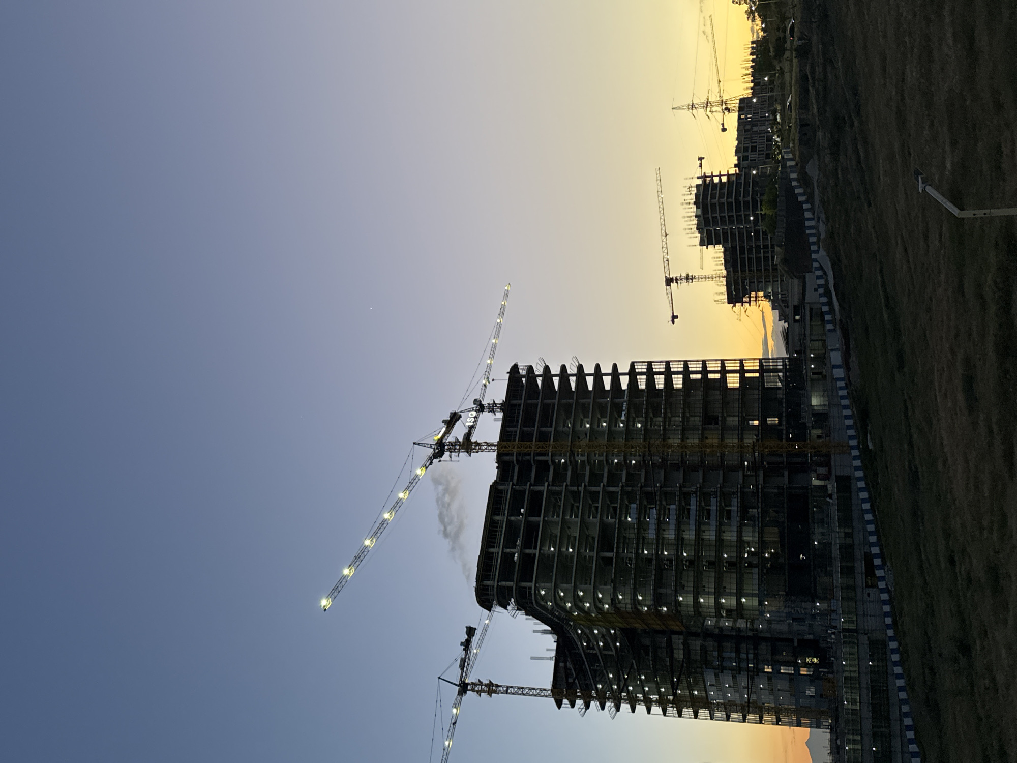 The image depicts a large construction site in Yerevan, Armenia, captured during sunset or dusk. A massive, multi-story building, still under construction, dominates the left side of the frame. Its concrete and steel skeleton is clearly visible, with some floors already fitted with windows and interior lights illuminating various sections.
Multiple tower cranes surround the main structure. One prominent crane, positioned behind and slightly above the primary building, has brightly lit jibs and clearly displays the text "ISG" on its side. Another large crane is visible near the bottom of the building, also illuminated. In the background, further to the right, additional buildings in various stages of construction can be seen, indicating extensive development.
The sky exhibits a striking gradient, transitioning from a deep, clear blue on the far left to a warm orange and yellow hue on the right, characteristic of a sunset. A faint, bright point, possibly a star or planet, is visible in the upper left portion of the blue sky. The right side of the image features undeveloped, grassy terrain with a path and some fencing, contrasting with the bustling construction activity. No people are visibly discernible in the scene.