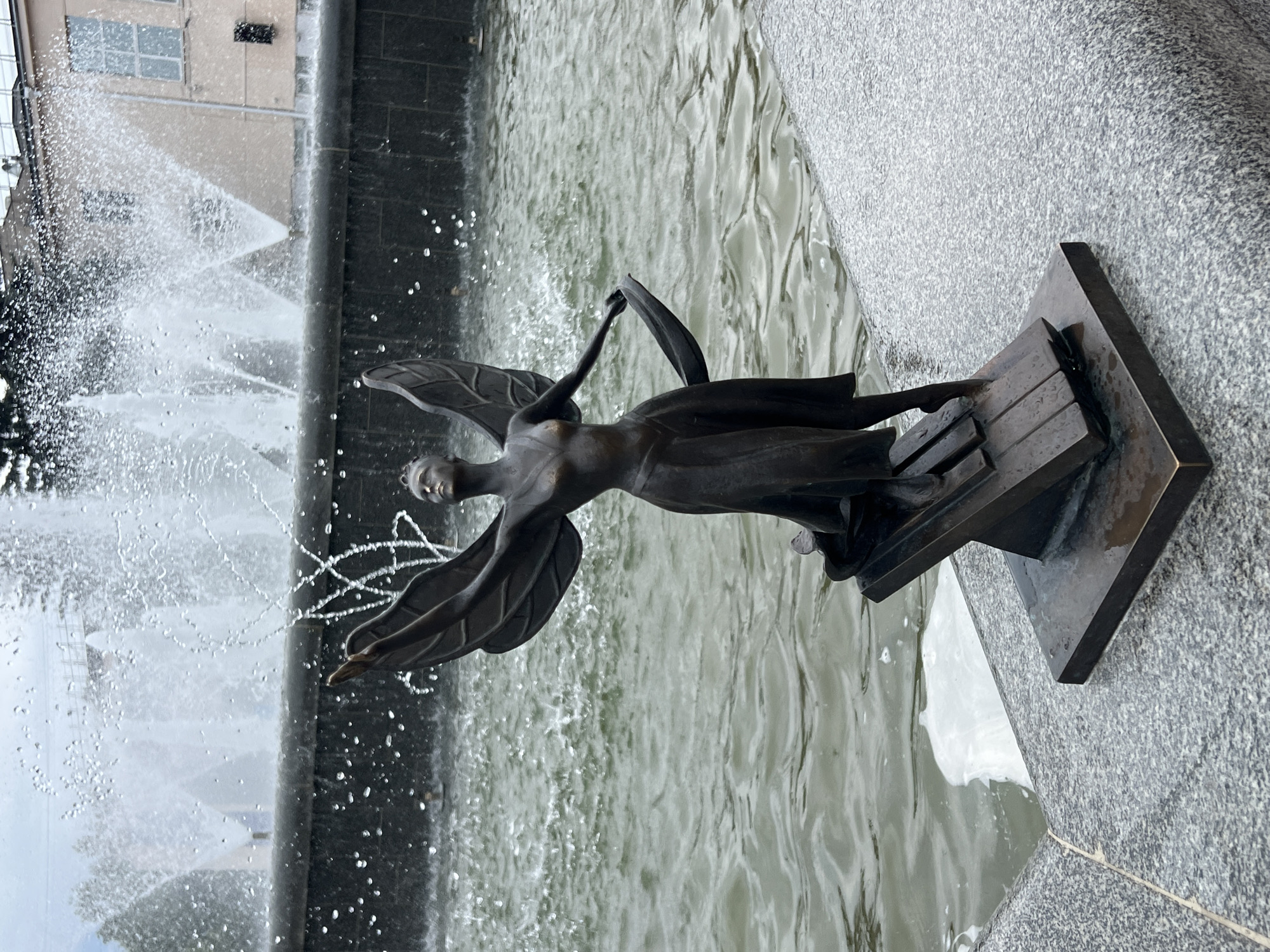 This is a bronze statue of a winged female figure with her arms outstretched and head tilted upwards, appearing to be in mid-dance. She is standing on a pedestal that resembles piano keys. The statue is positioned on the edge of a fountain with water spraying upwards and falling into the basin. The background shows a building and trees, and the overall environment appears to be outdoors in a public space. The statue is located in the city of Chaikovsky, Russia.